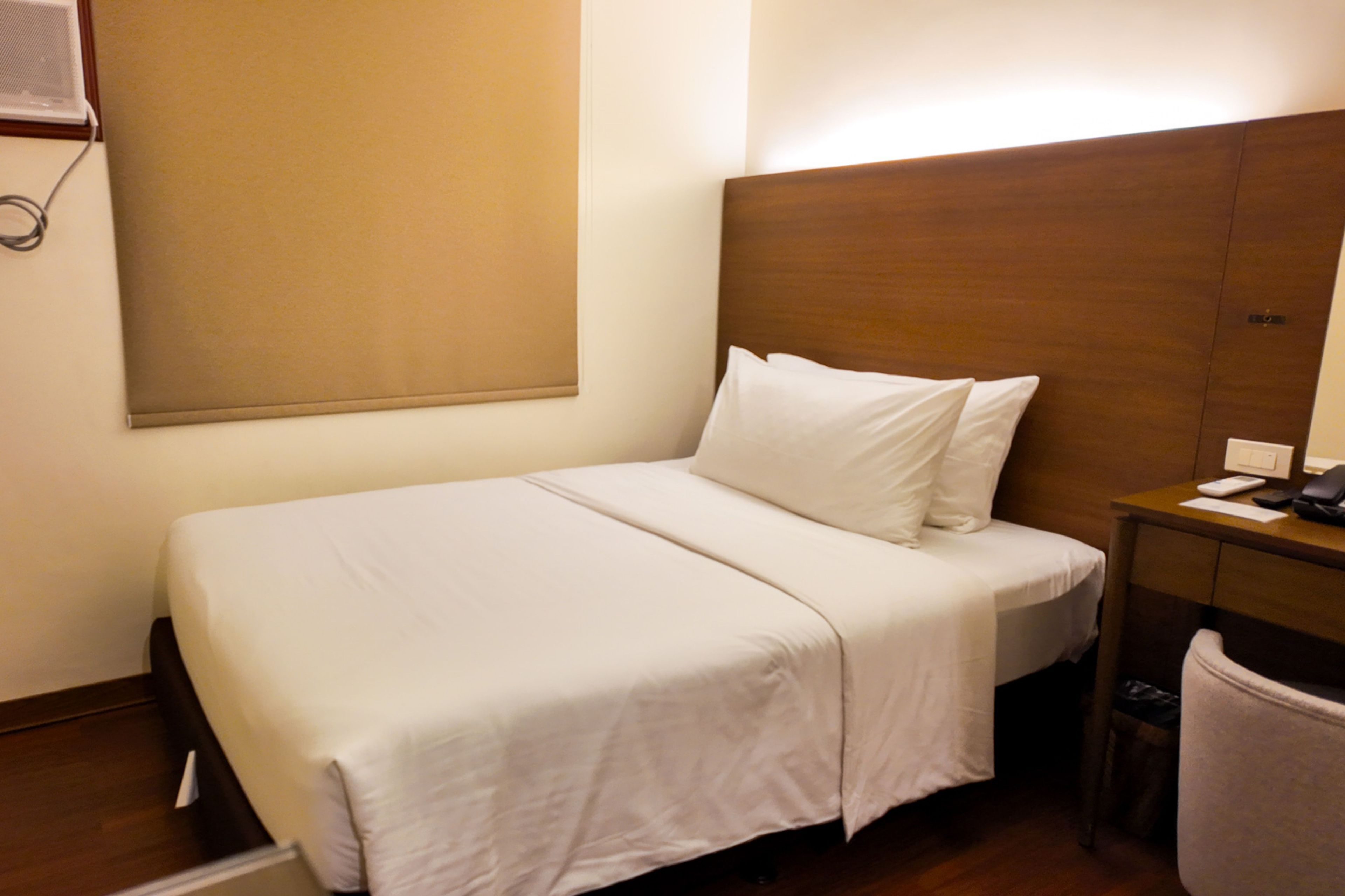 Habitación de hotel con una cama individual, cabecera de madera, mesita de noche con teléfono y una silla.