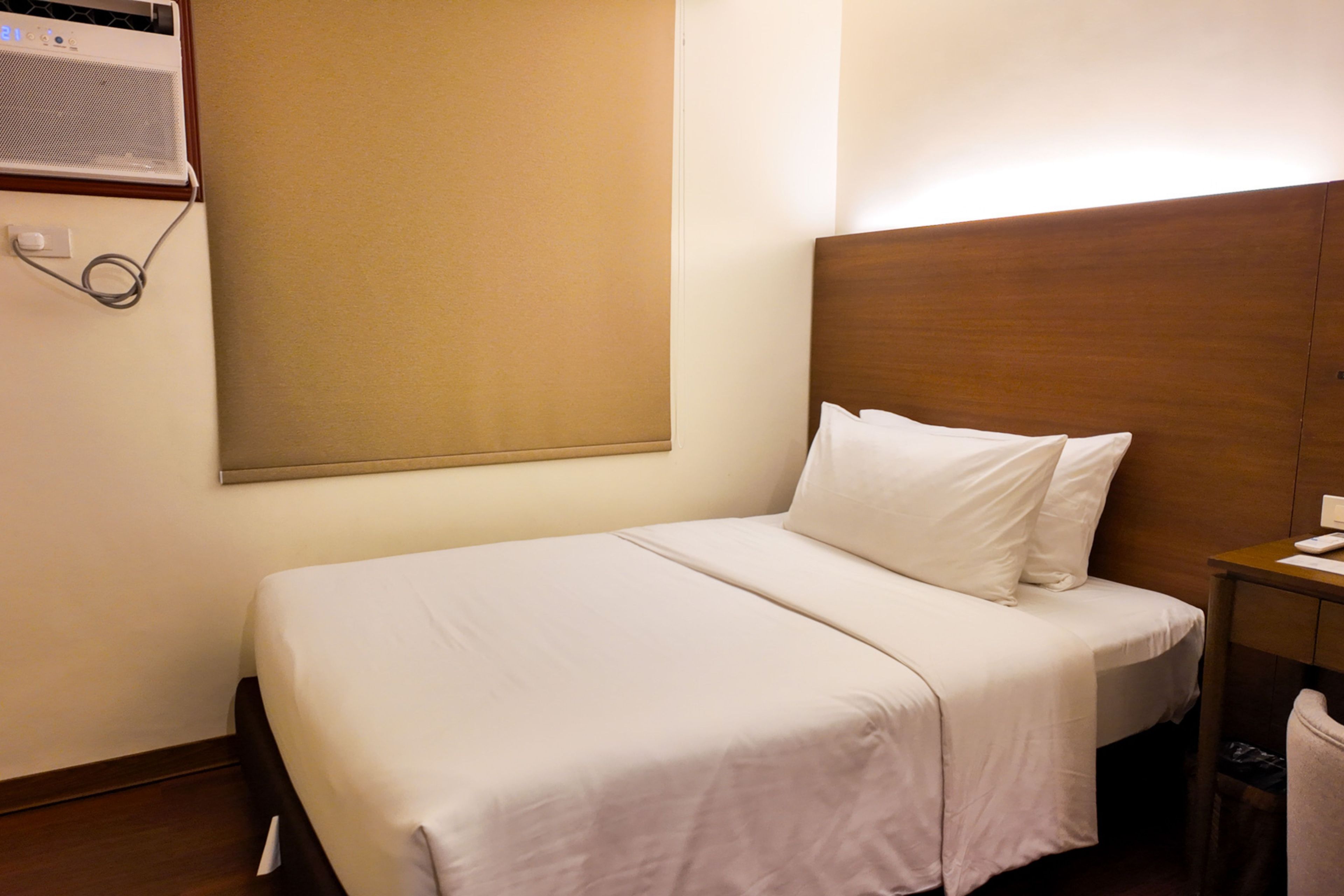 A hotel room with a single bed, a wooden headboard, a bedside table with a lamp, and a wall-mounted air conditioning unit.