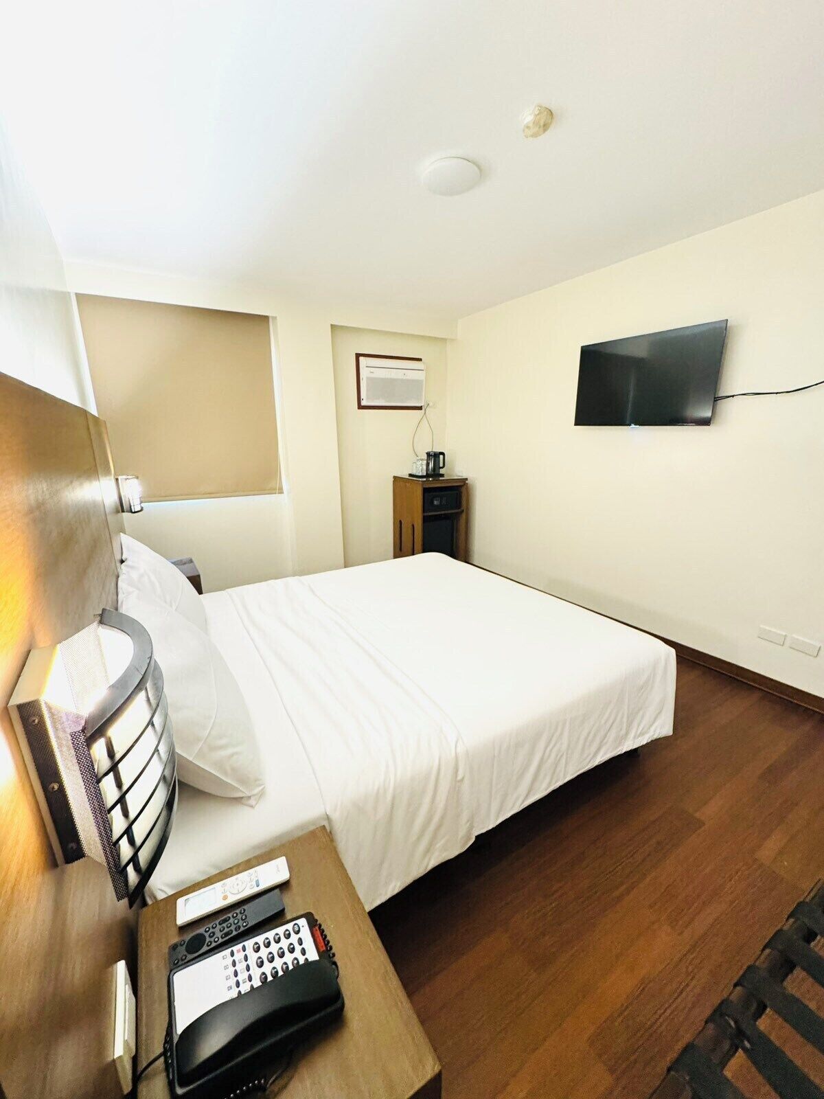 A hotel room with a bed, a bedside table with a phone, a TV mounted on the wall, and a small cabinet with an air conditioner.