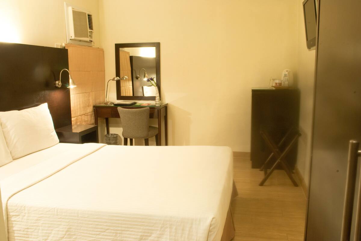 Habitación de hotel con cama, escritorio, silla, espejo, armario y refrigerador.
