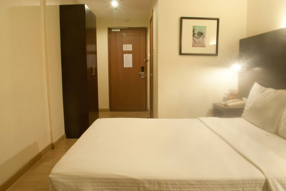 Habitación de hotel con una cama, una mesita de noche, un cuadro enmarcado en la pared y una puerta que da a otra habitación.