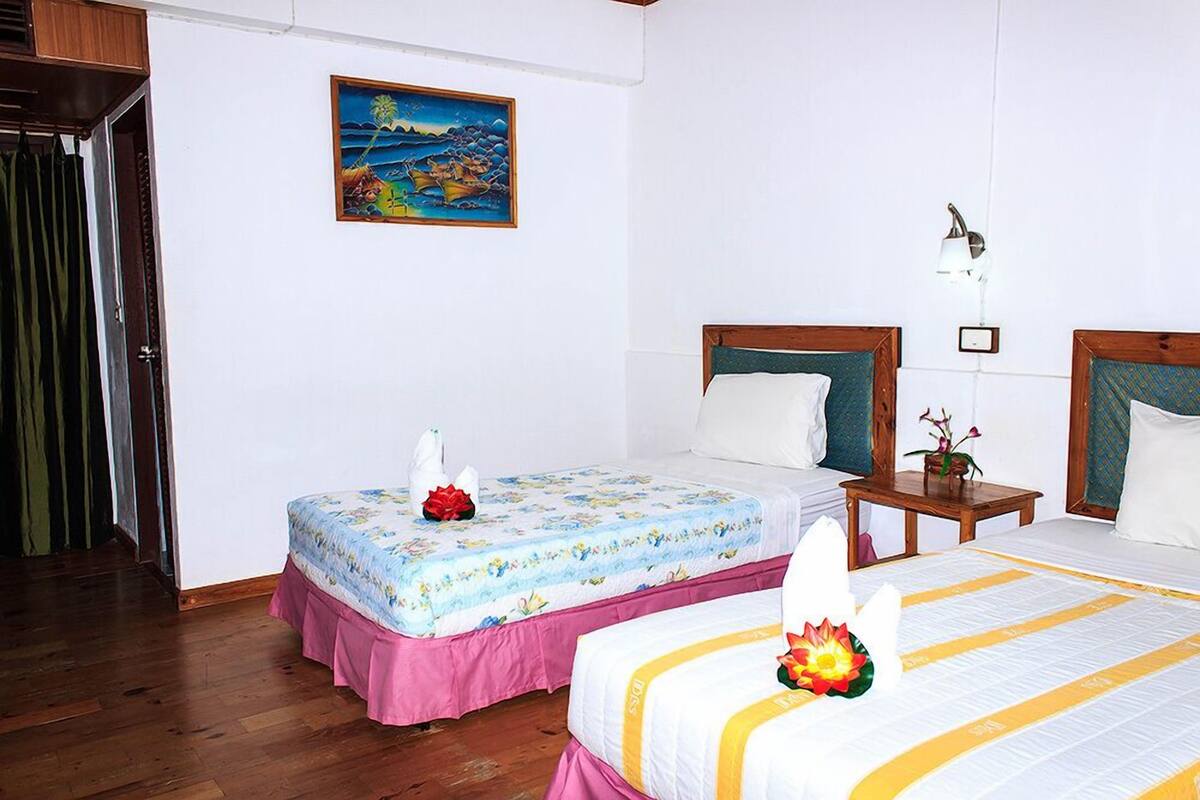 A hotel room with two beds, a wooden floor, a painting on the wall, and a nightstand with a lamp.
