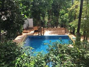 A rectangular swimming pool with a tiled edge, surrounded by lush greenery and trees. There are wooden lounge chairs and a small white building in the background. - Reuan Thai Village Samui (Koh Samui)