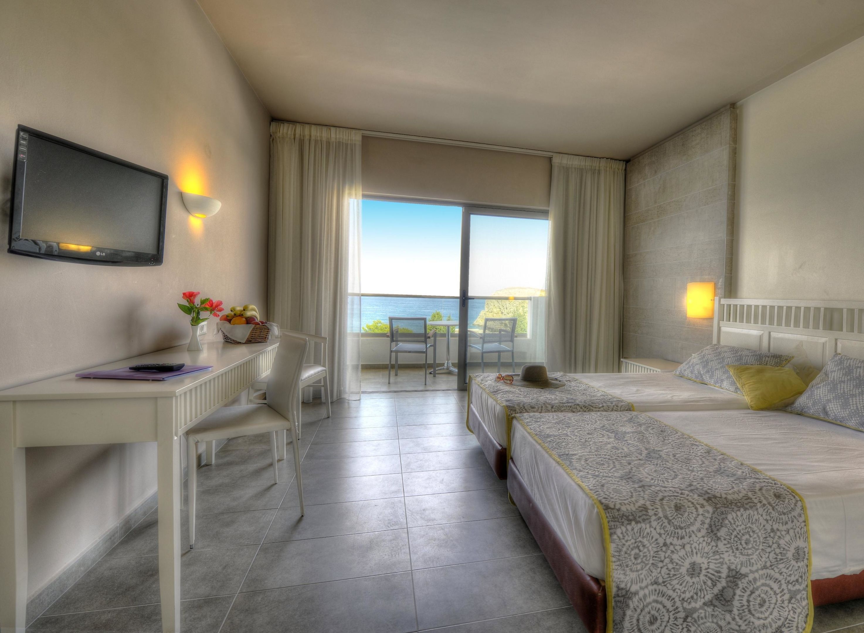 A hotel room with a bed, a desk, a chair, a TV, and a balcony with a view of the sea.
