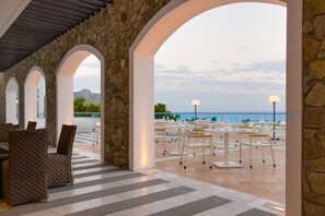 An outdoor dining area with stone arches, white tables, and chairs, overlooking a body of water and mountains. - Porto Angeli Beach Resort – All Inclusive (Rhodes)