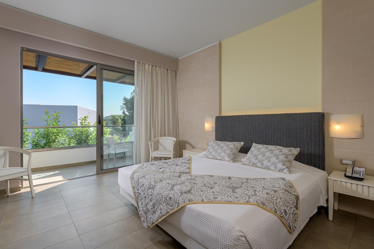 A modern bedroom with a large bed, a chair, a bedside table, and a view of greenery through a sliding glass door.