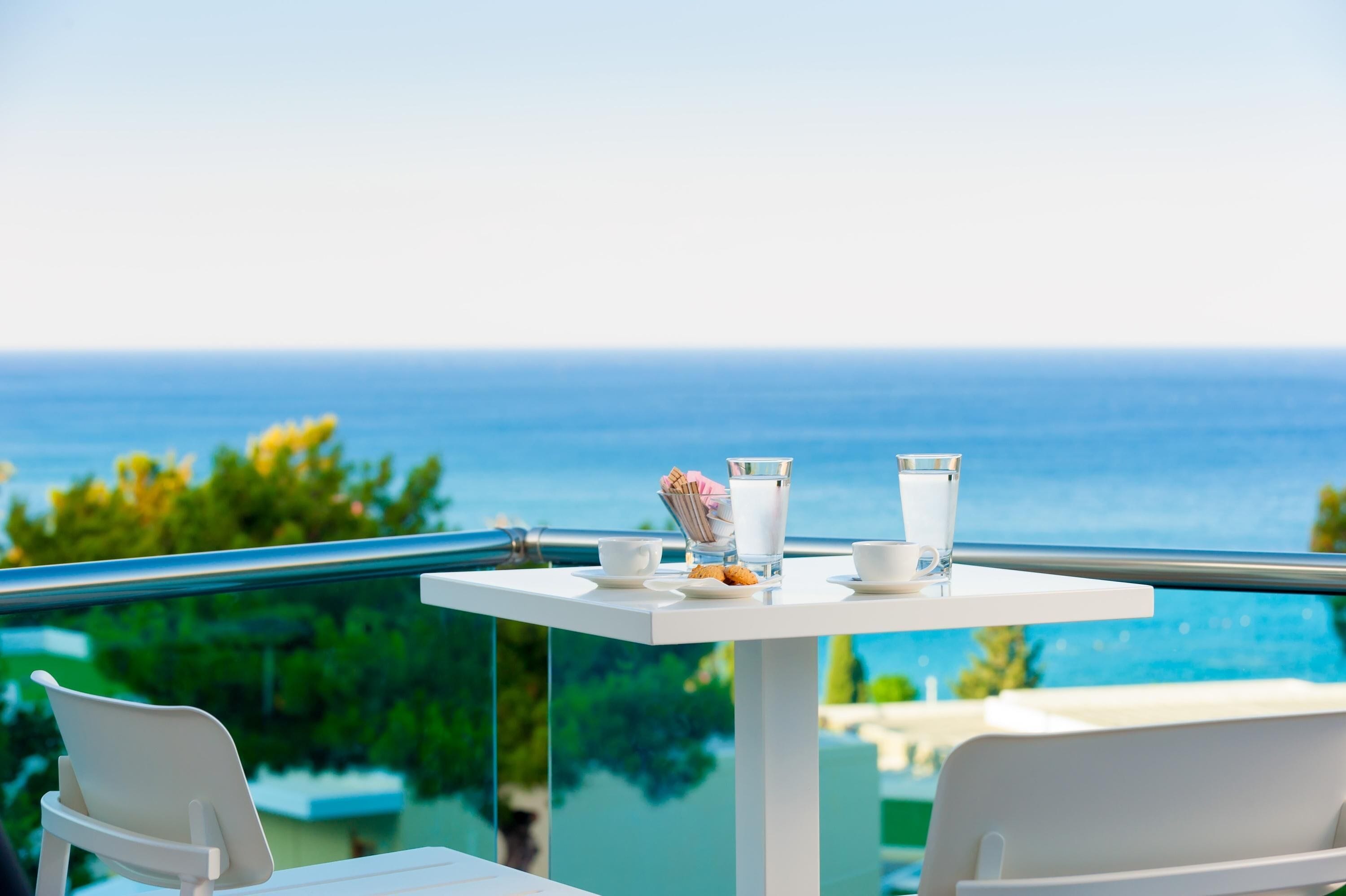 A white table with a glass of water, a cup, and a plate of cookies, overlooking a pool and the ocean.