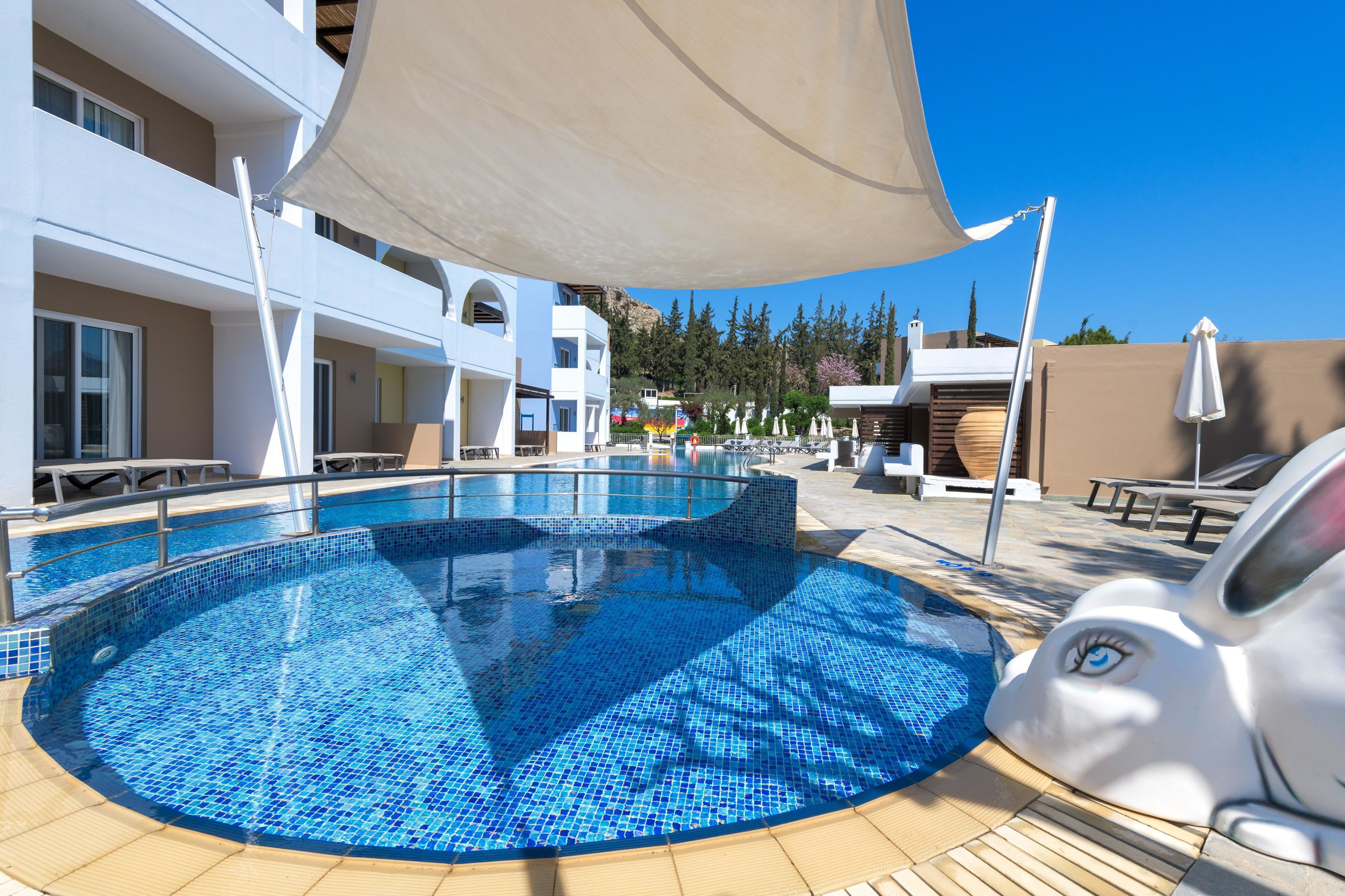 Una piscina con mosaicos azules, rodeada de sillas de descanso y sombrillas, con edificios al fondo.