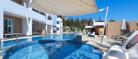 Una piscina con mosaicos azules, rodeada de sillas de descanso y sombrillas, con edificios al fondo.
