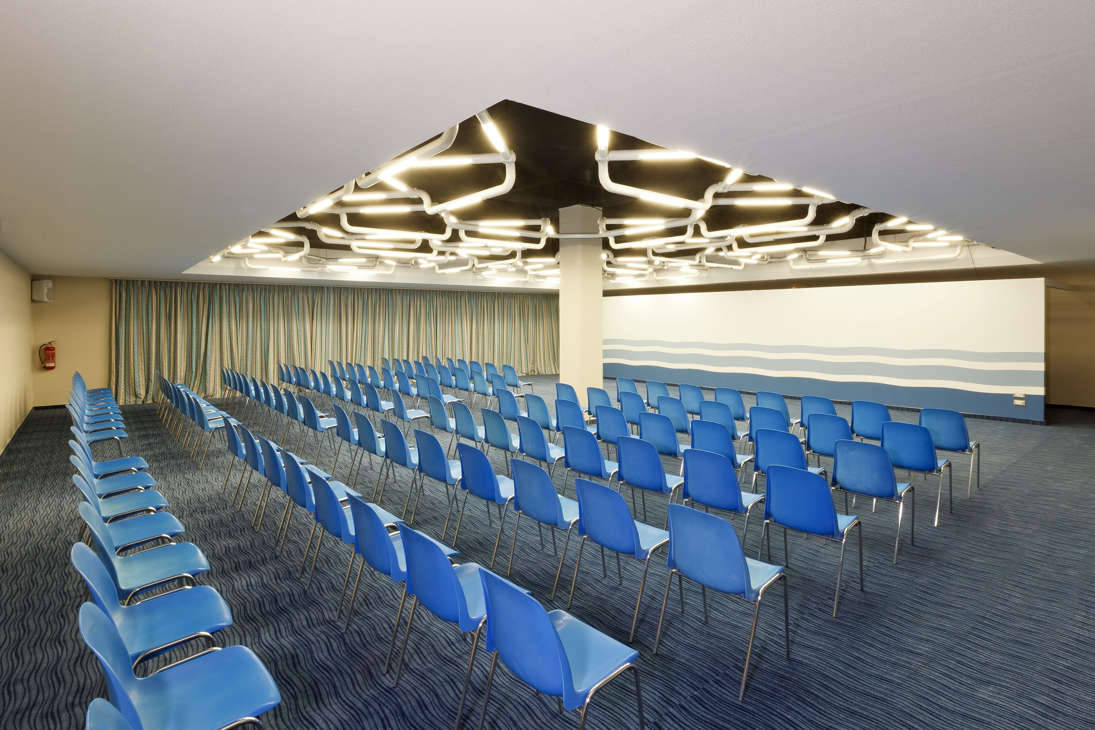 Un salón de conferencias con filas de sillas azules, una alfombra estampada y un techo con una lámpara de diseño.