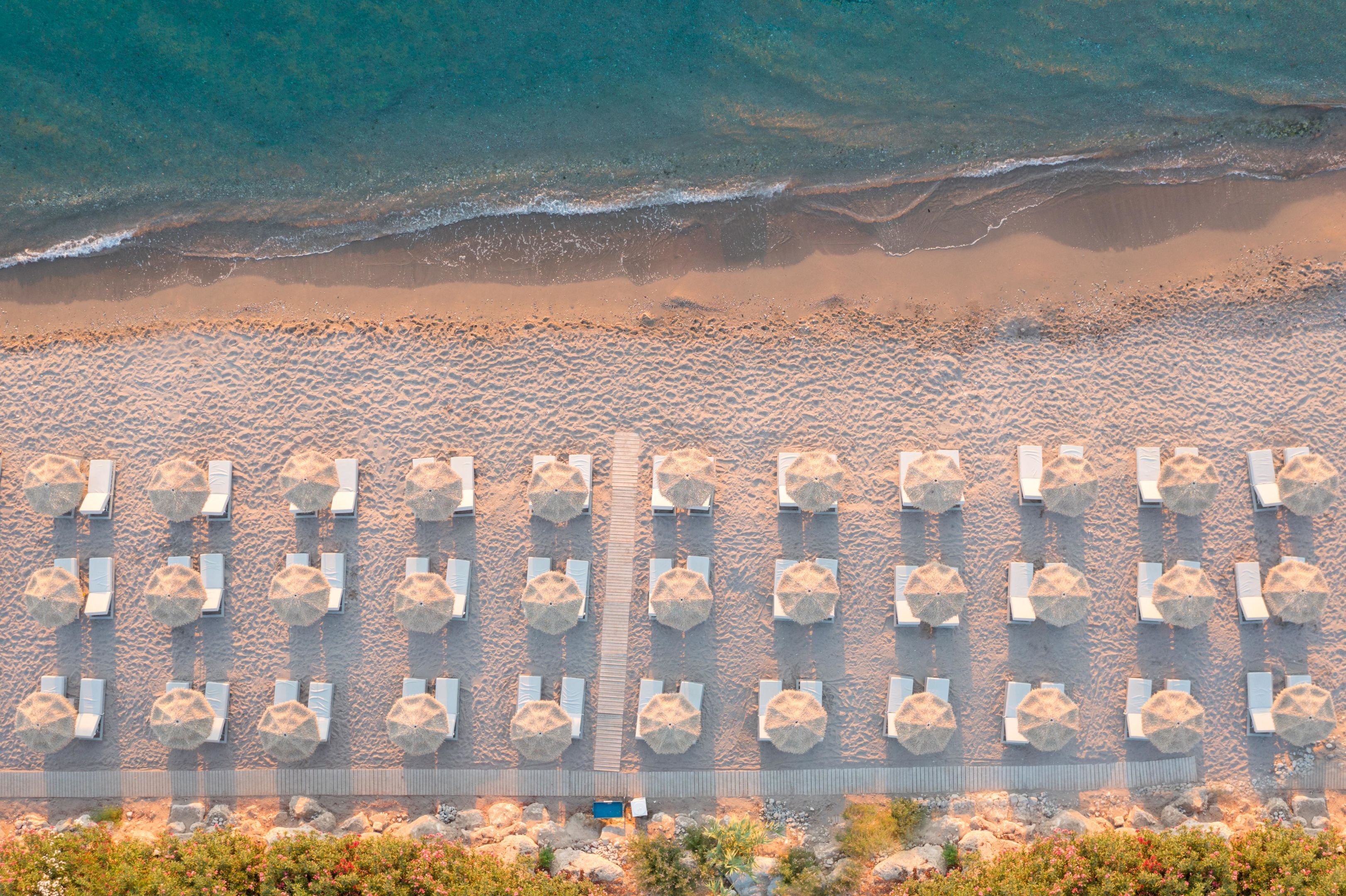 Une vue aérienne d’une plage bordée de rangées de chaises longues et de parasols, d’une côte rocheuse et offrant une vue dégagée sur l’océan.