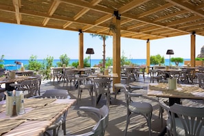 An outdoor dining area with tables and chairs, a view of the sea, and a thatched roof structure. - Porto Angeli Beach Resort – All Inclusive (Rhodes)
