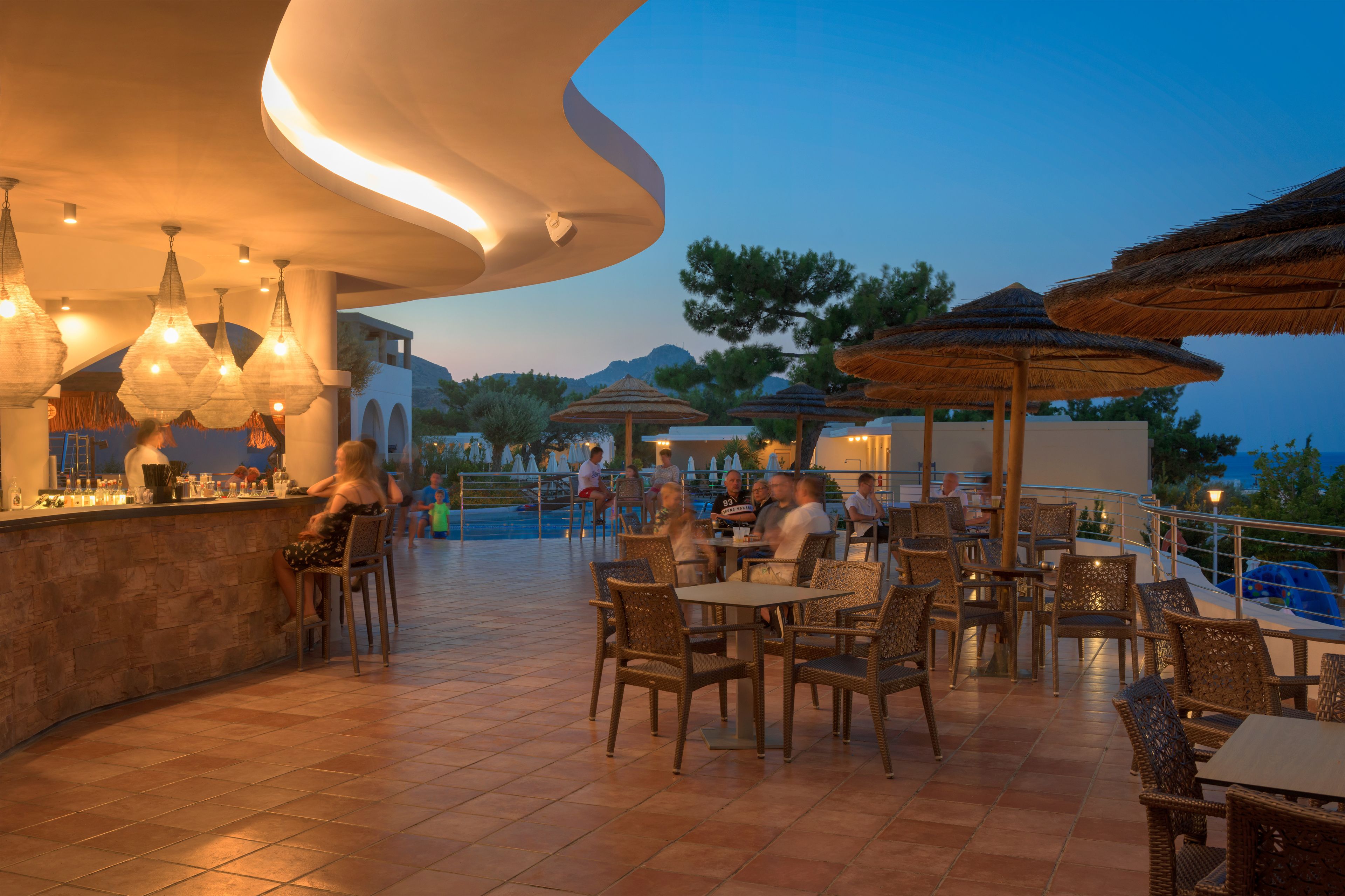 An outdoor dining area with wicker chairs and tables, a bar with a stone counter, and thatched umbrellas.