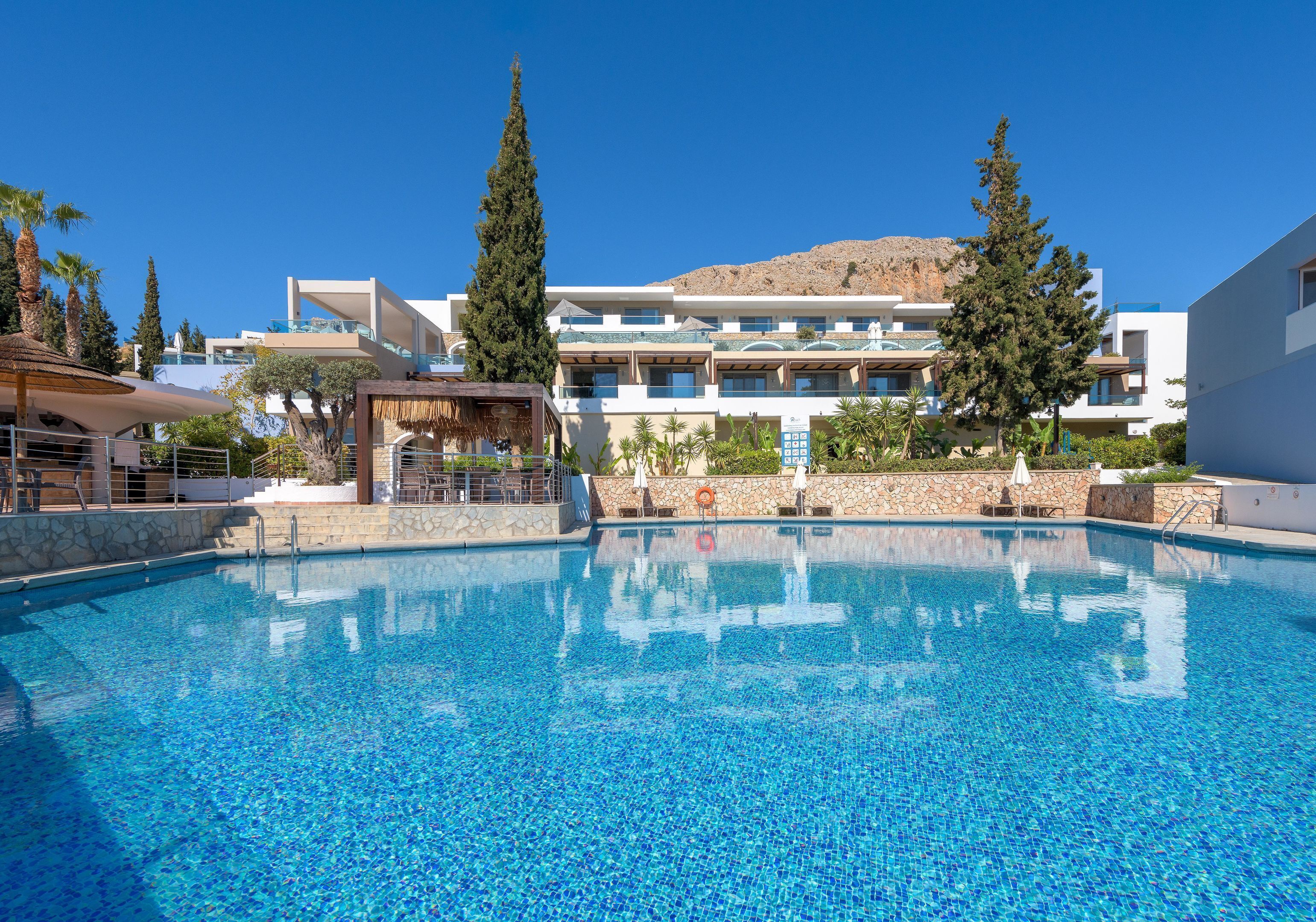 A swimming pool with a clear blue water, surrounded by a modern building with balconies and a mountain in the background.