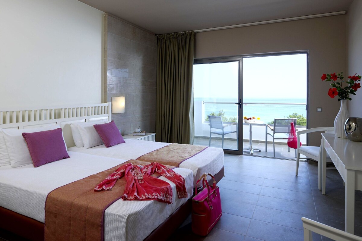 A hotel room with a bed, a desk, a chair, and a balcony with a view of the sea.