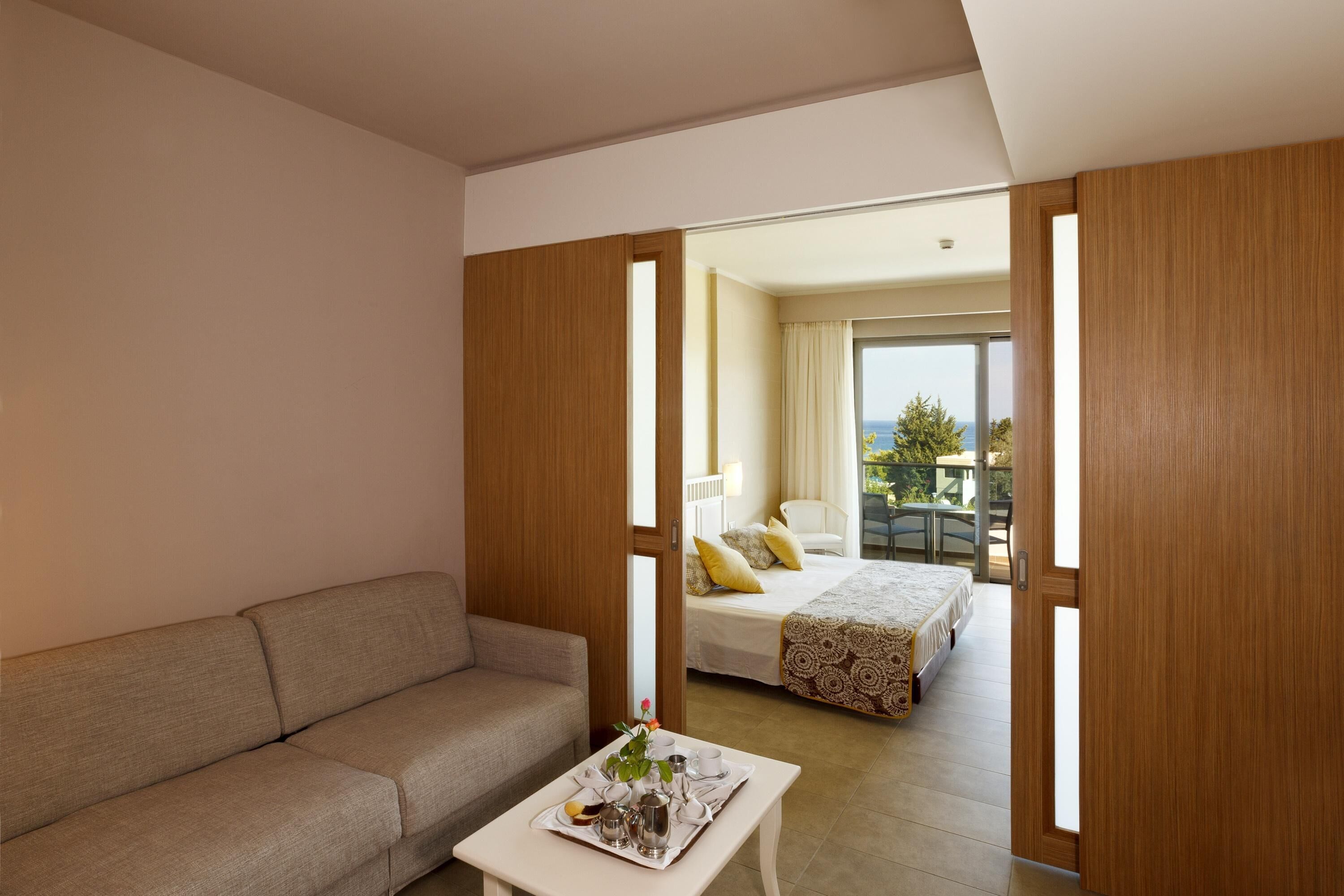 A modern hotel room with a sofa, a dining table with a tray of breakfast items, and a bed with a patterned bedspread.