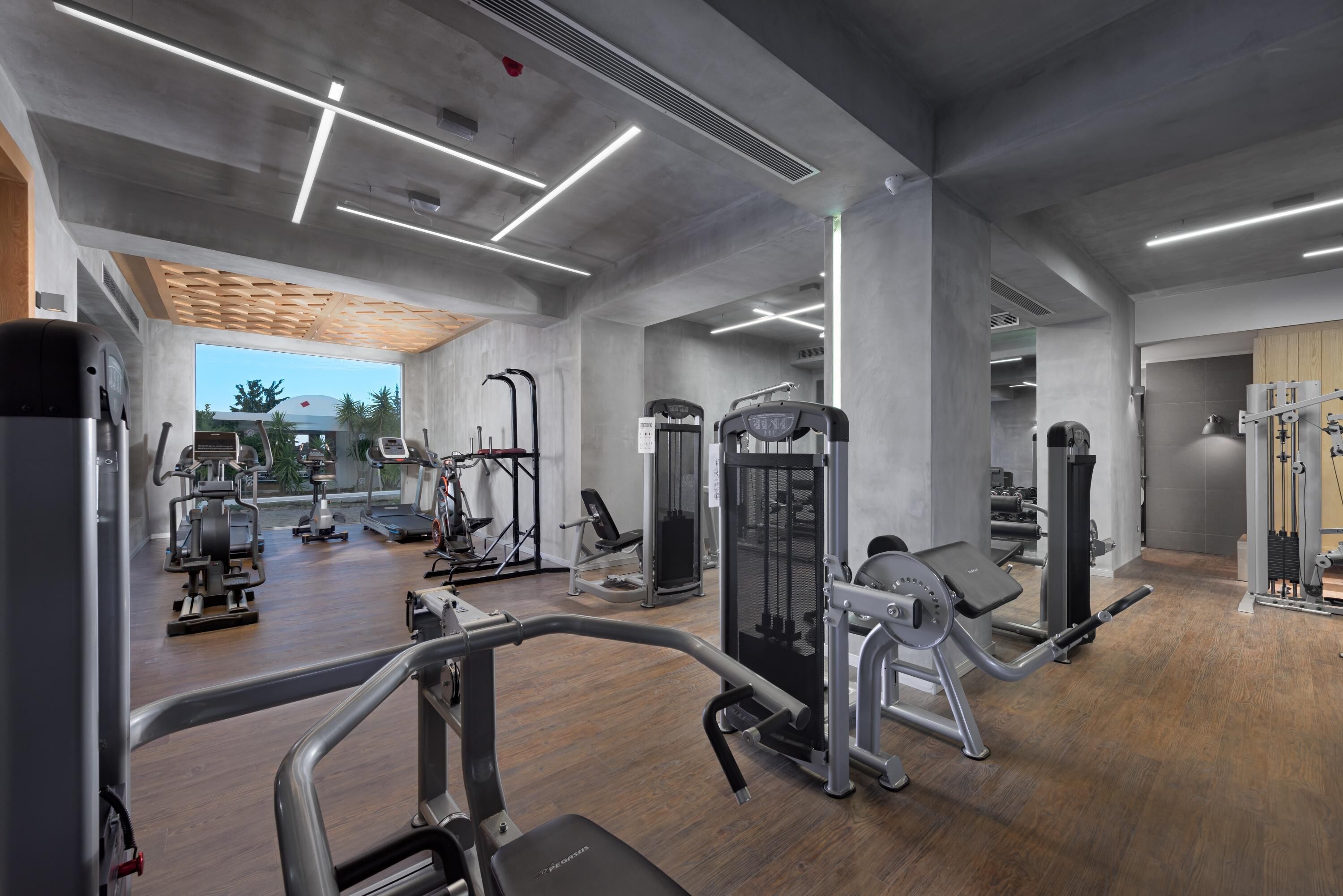A modern gym with various exercise equipment, large windows, and a wooden ceiling design.