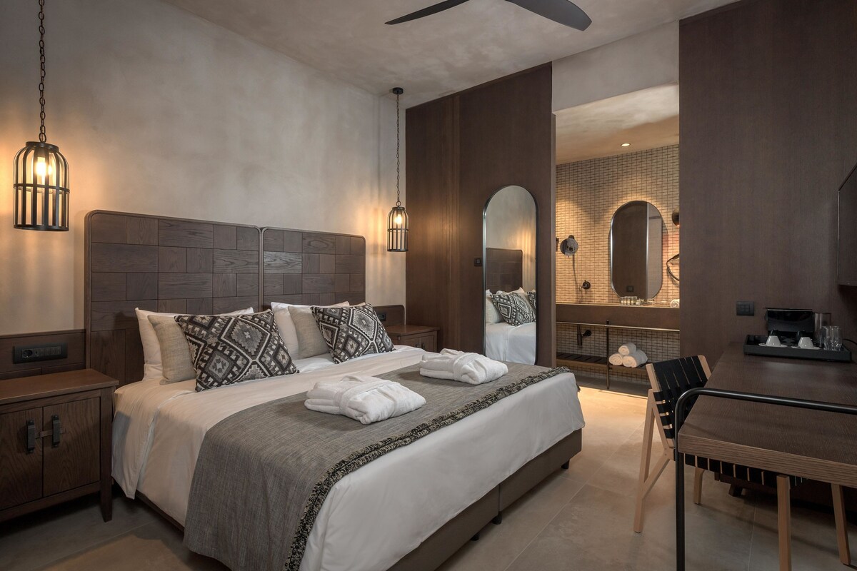 A modern hotel room with a large bed, bedside tables, a dining area, and a bathroom with a mirror and shower.