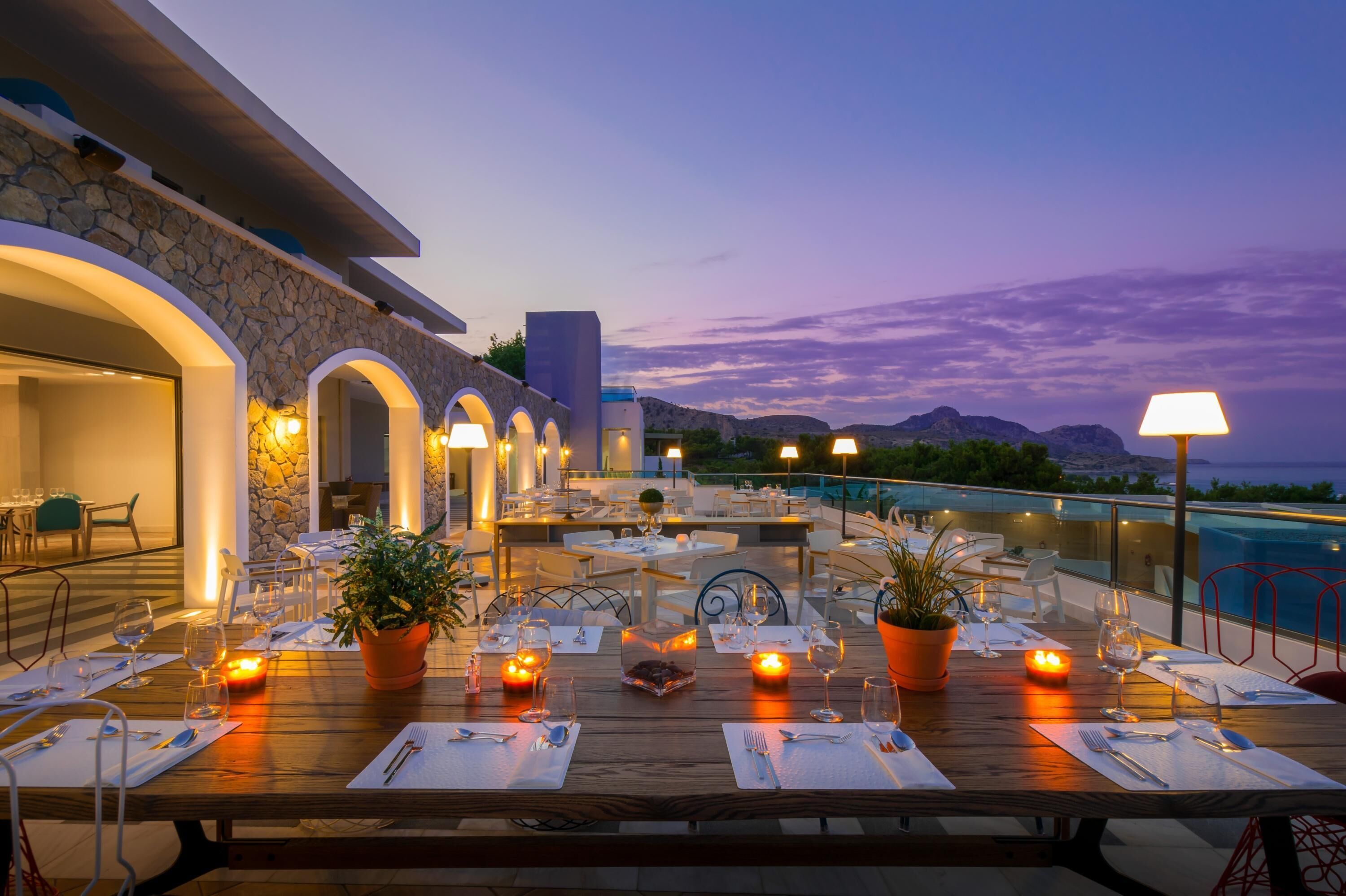 Un área de comedor en la azotea con mesas dispuestas para una comida, iluminación exterior y vistas a las montañas.