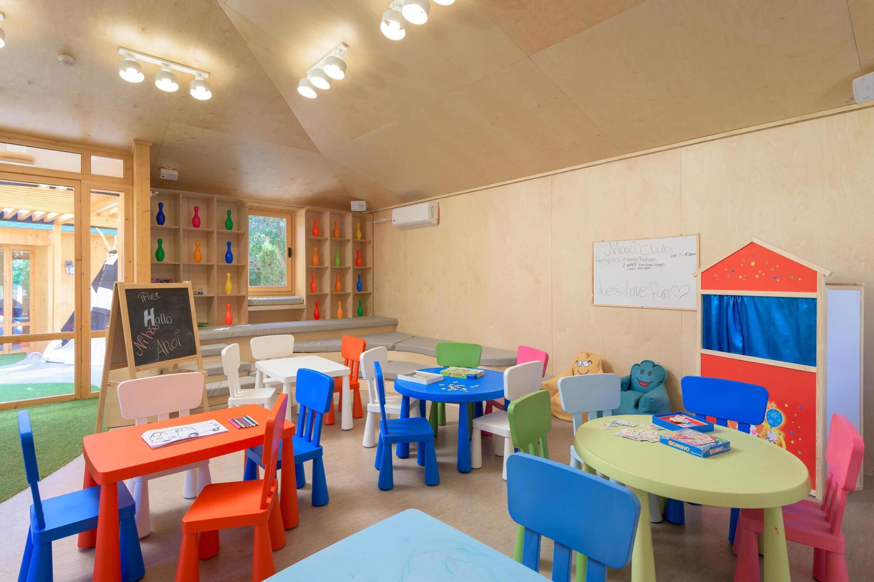 A children's playroom with colorful chairs and tables, a chalkboard, and a playhouse model.