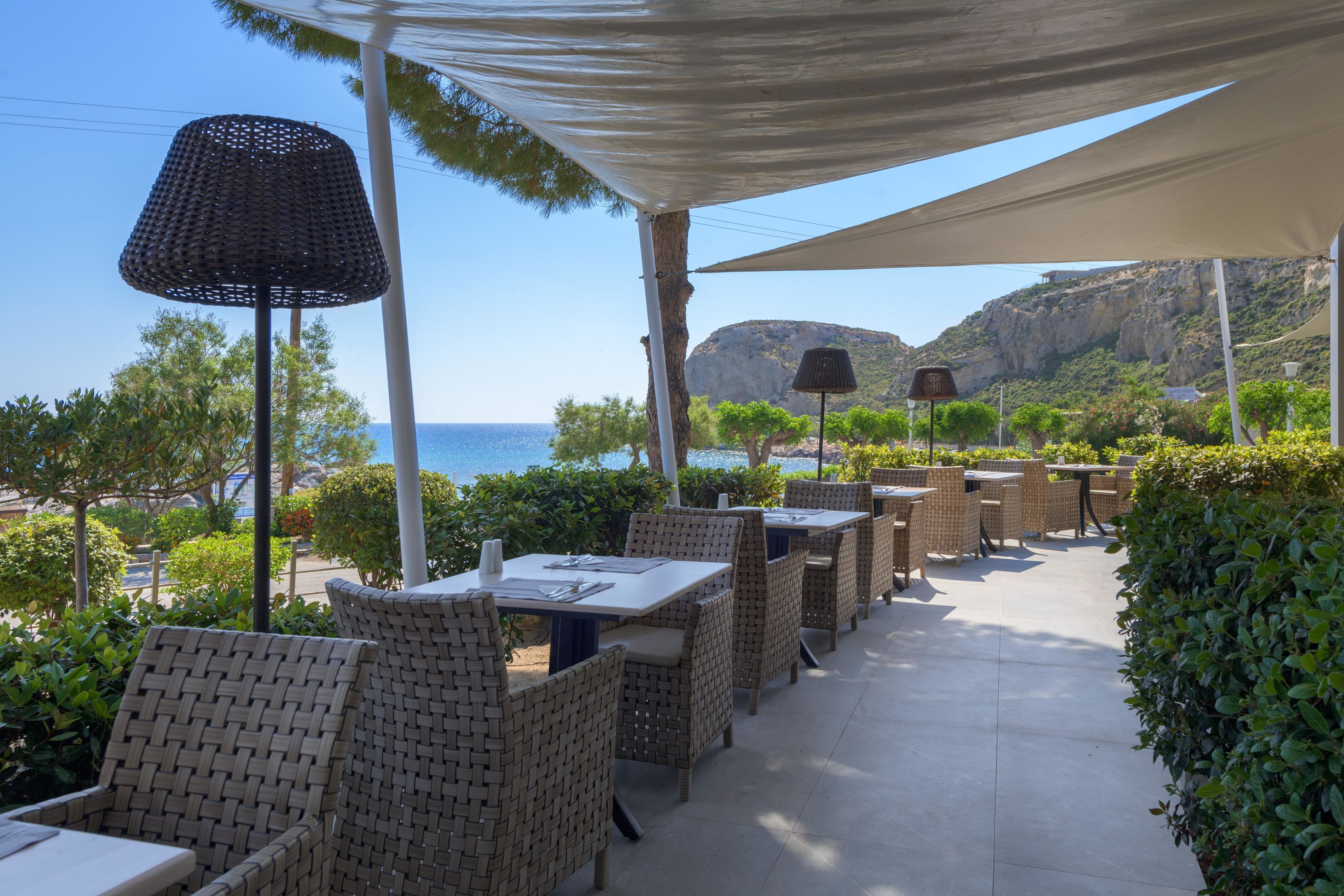 An outdoor patio with wicker furniture and tables, overlooking a coastal area with a view of the sea and hills.