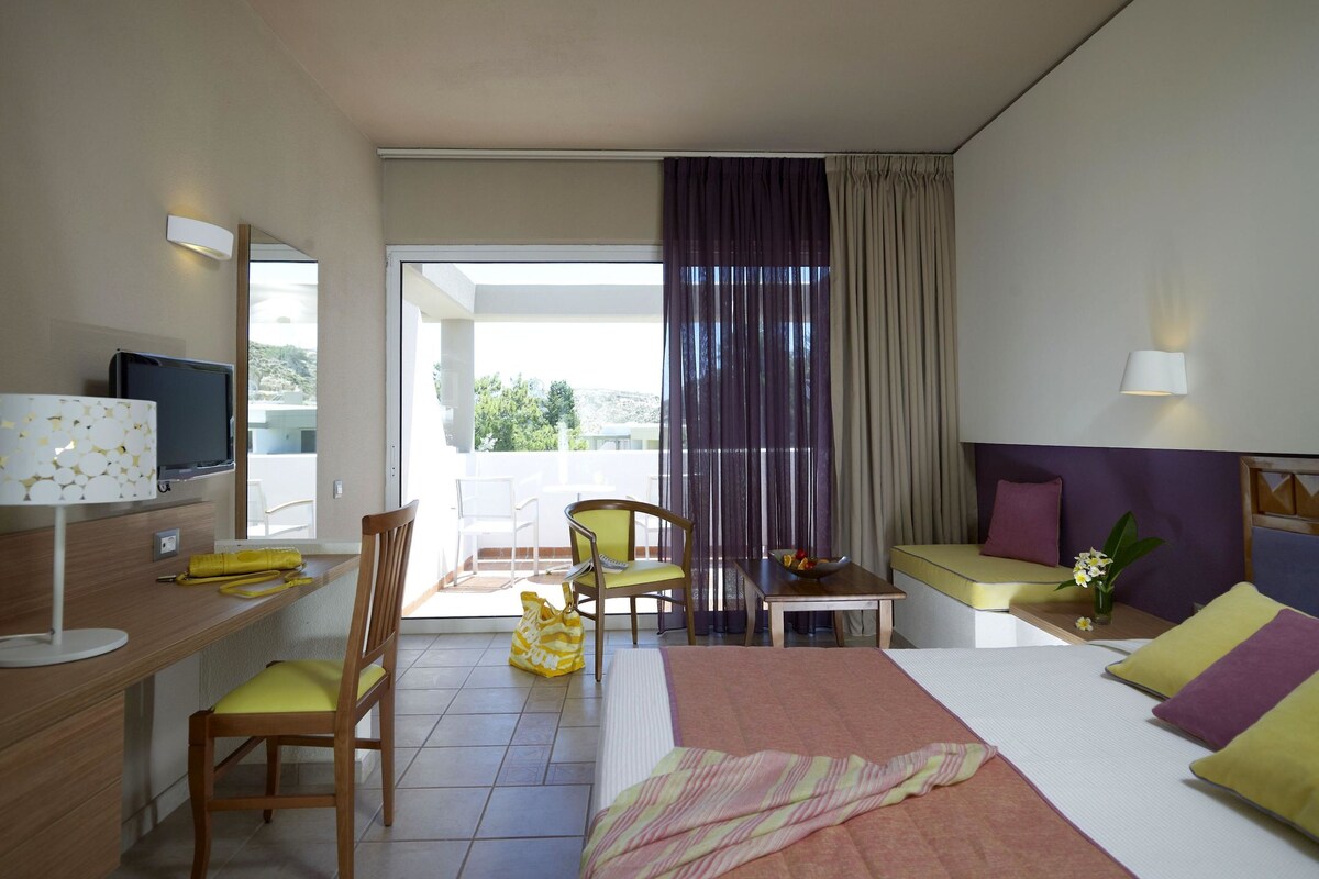 A hotel room with a bed, a desk, a chair, a small table, and a balcony with a view.