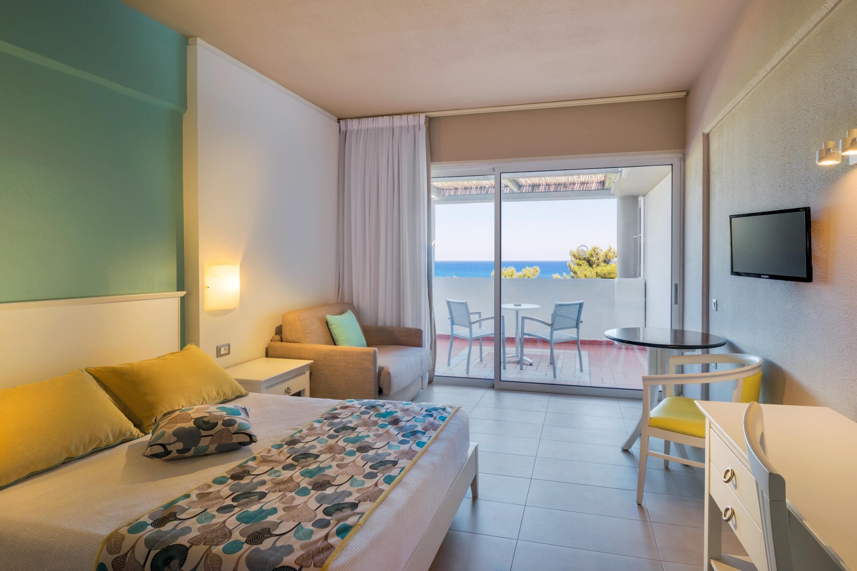 A hotel room with a bed, a desk, a chair, a TV, and a balcony with a view of the sea.