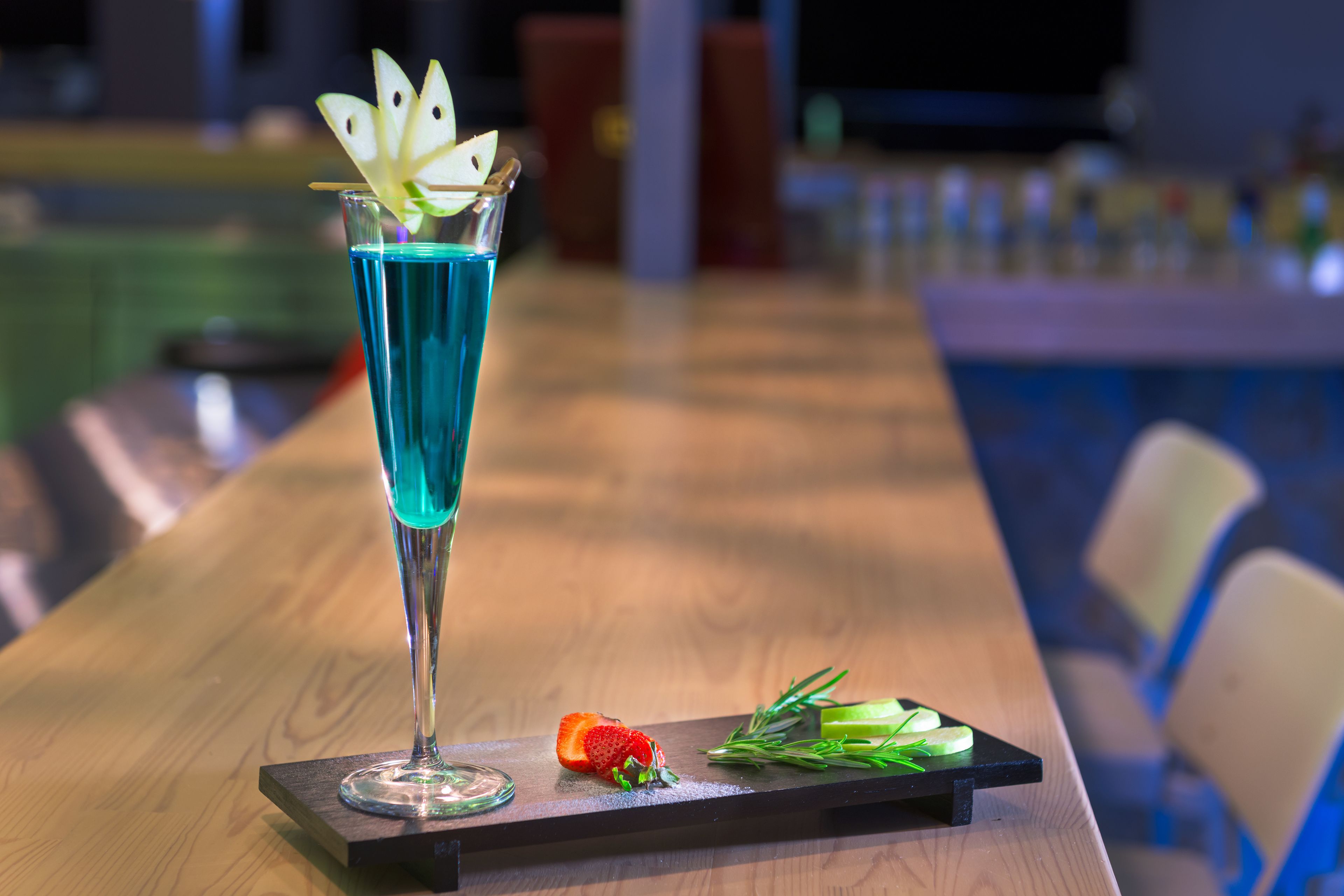 A cocktail glass with a blue liquid, a strawberry slice, and a sprig of rosemary on a wooden tray.