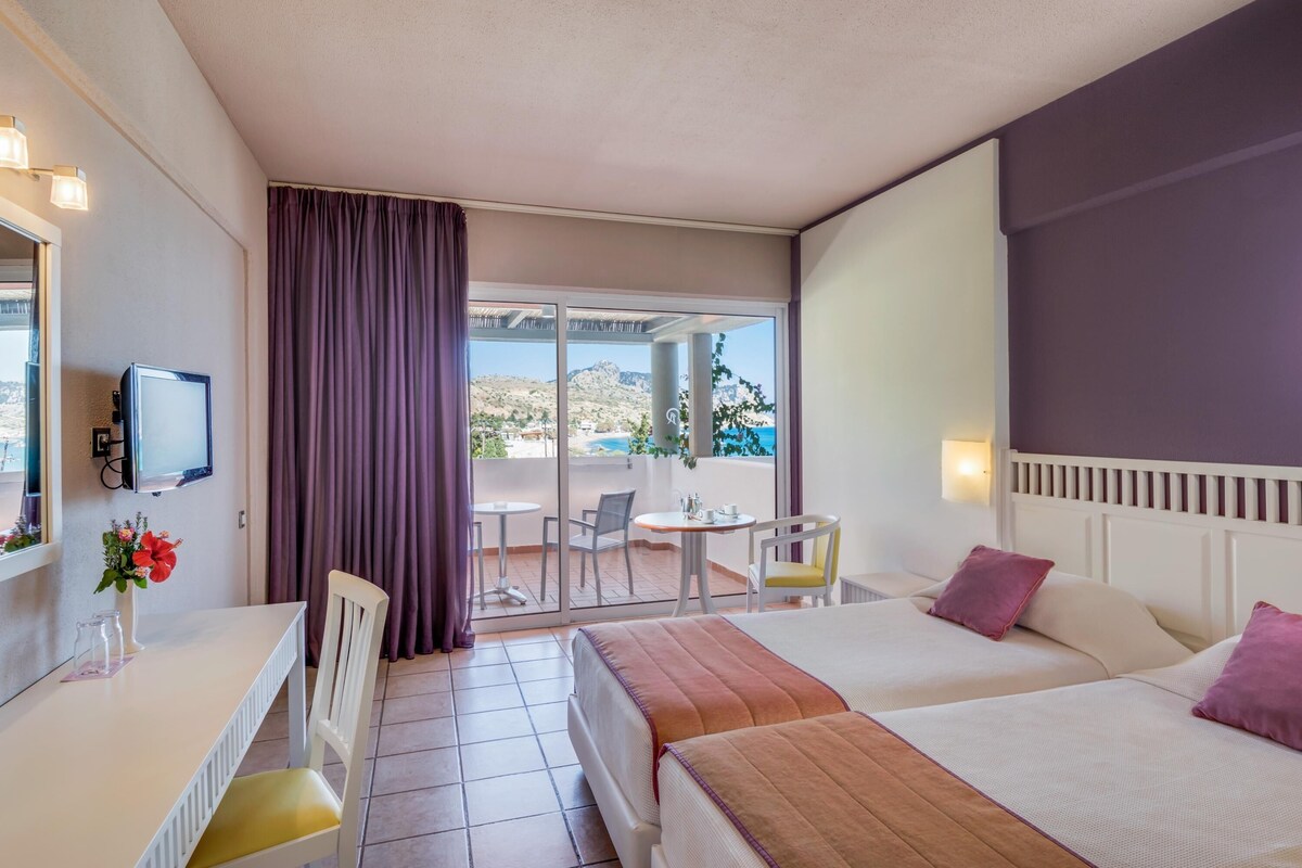 A hotel room with two beds, a desk with a TV, a small table, and a view of a mountainous landscape through a sliding glass door.