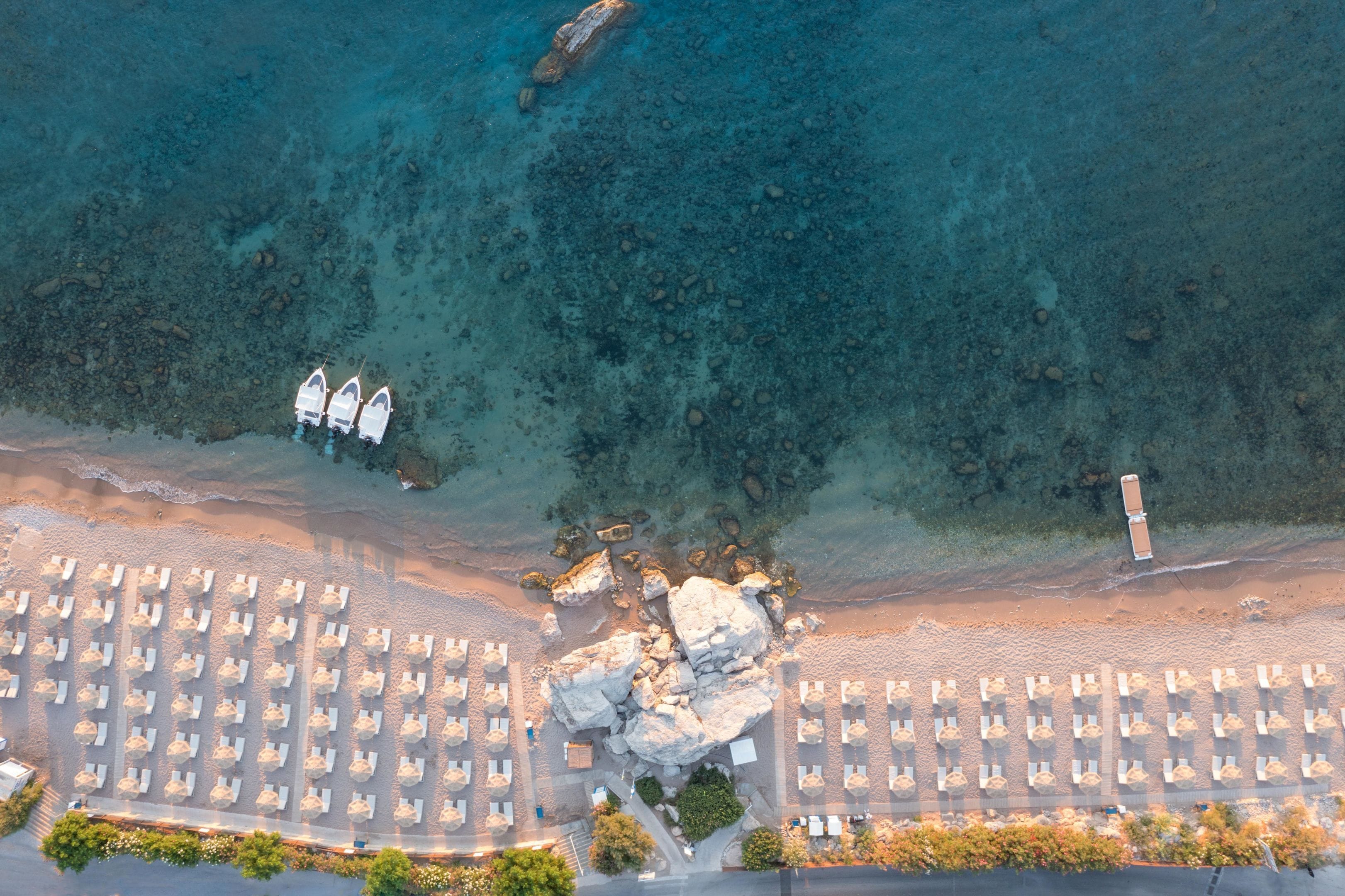An aerial view of a beach with rows of sunbeds, a rocky area, and boats in the water.