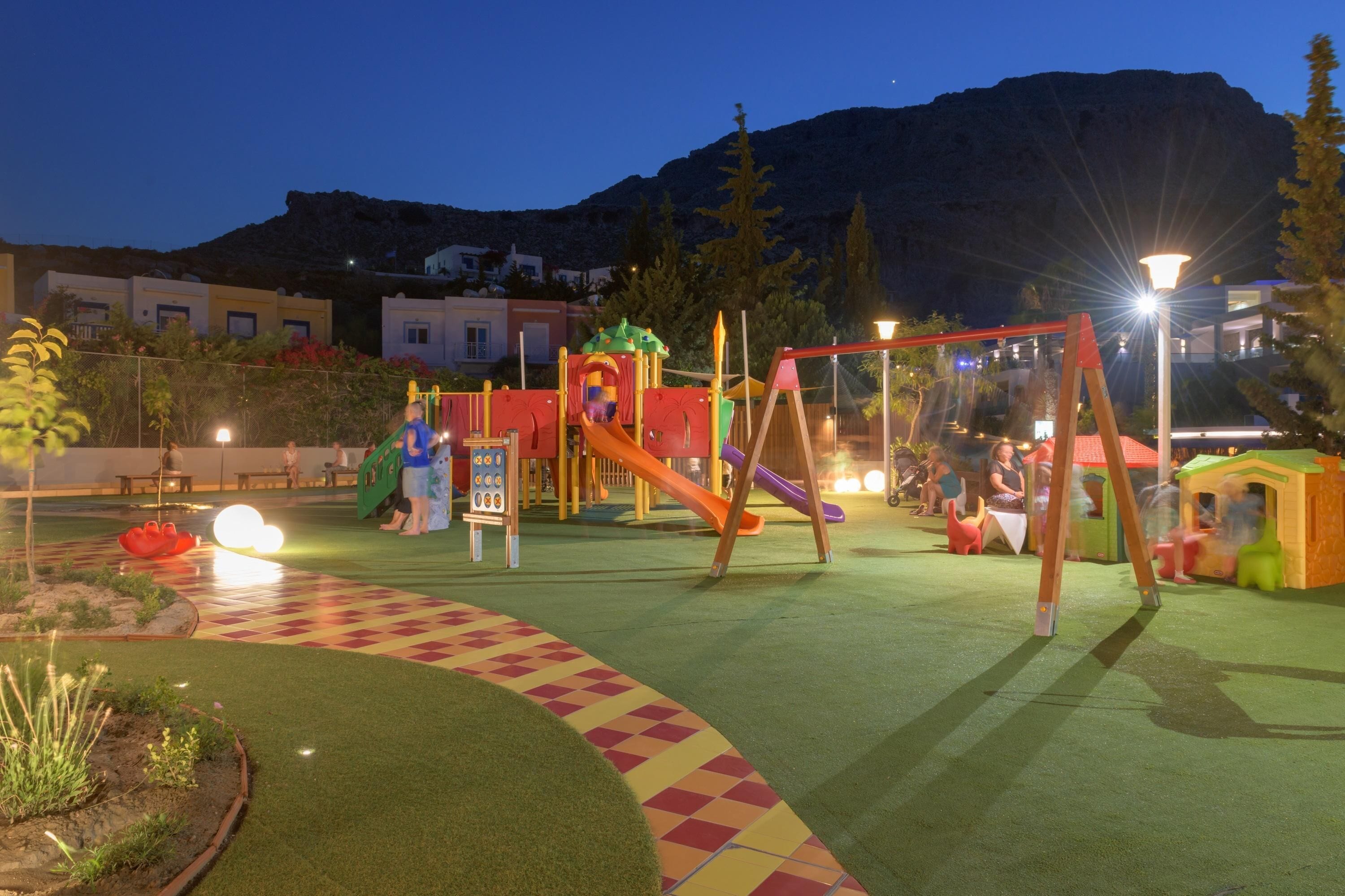 A playground with swings and slides, surrounded by artificial grass and a checkered path.