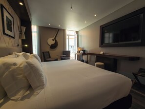 Et hotelværelse med en stor seng, et tv ophængt på væggen, en guitar og et lille bord med en lampe.