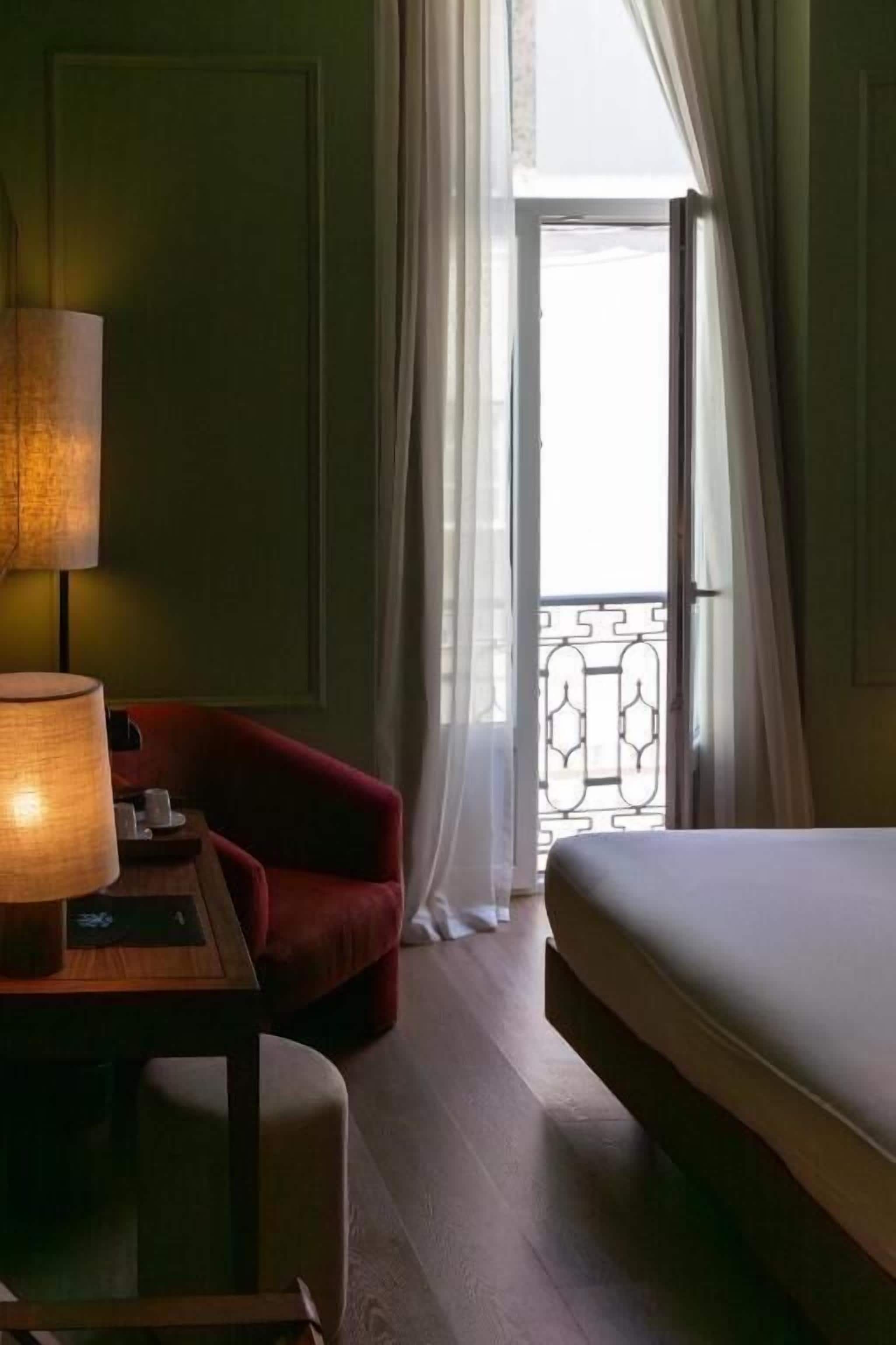 Ein Hotelzimmer mit einem Bett, einem Schreibtisch mit Lampe, einem roten Sessel und Blick durch ein mit Vorhängen drapiertes Fenster hinaus.