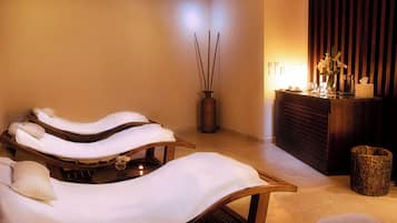 A spa room with three massage tables, a wooden cabinet with a vase of flowers, and a decorative vase with sticks.