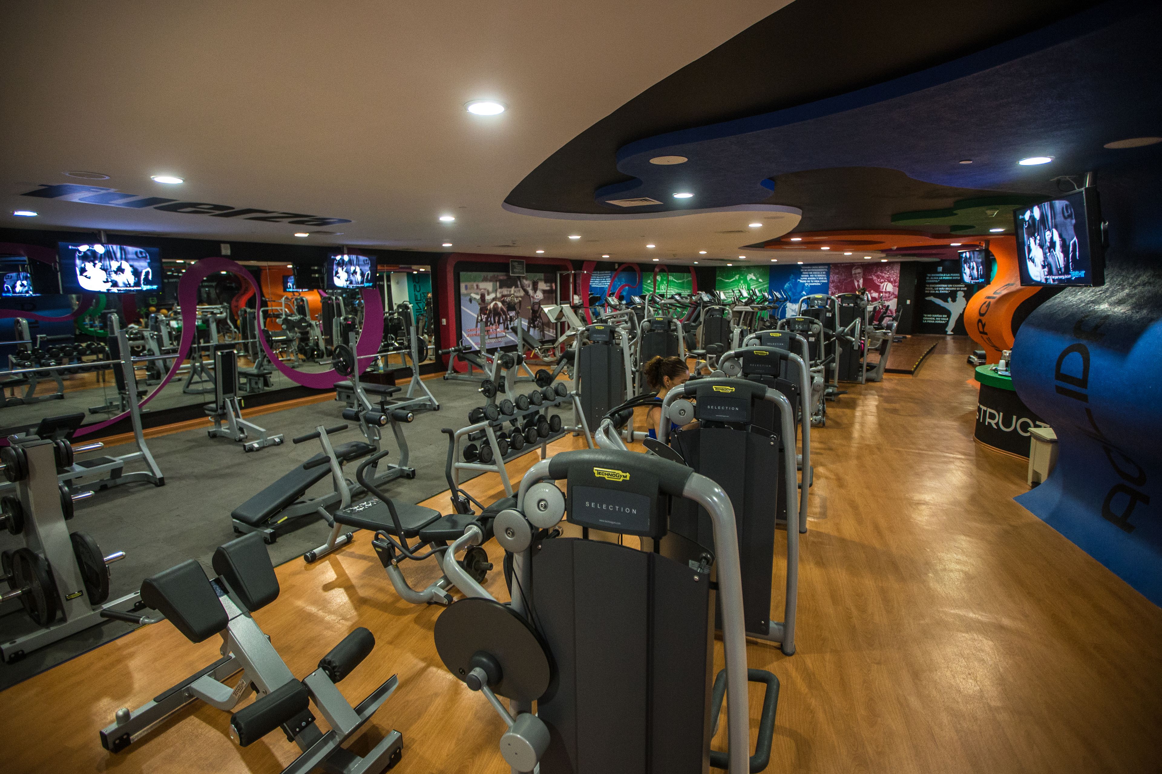 A gym with various exercise machines and weights, wooden flooring, and multiple TV screens displaying workout content.