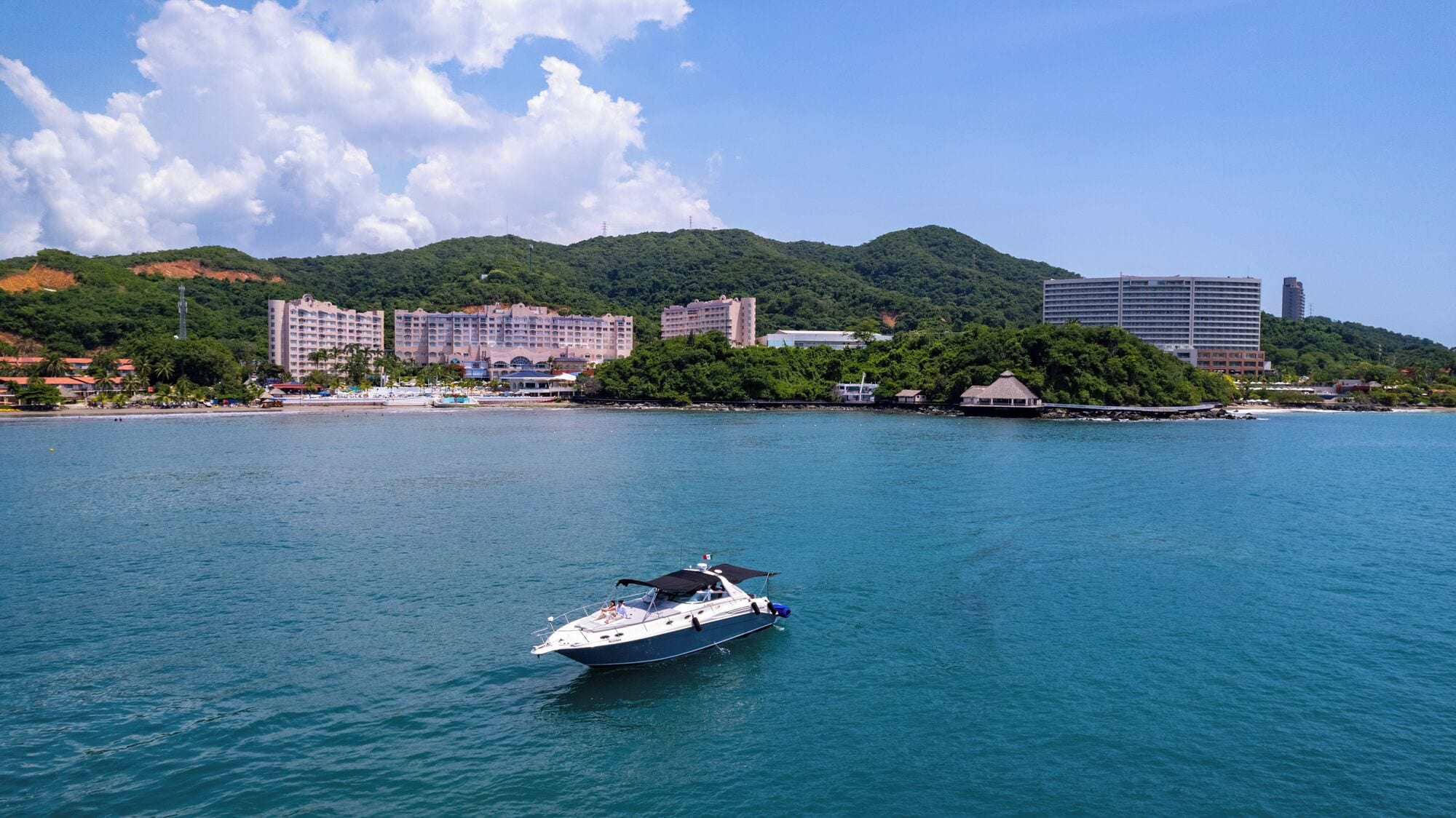 A coastal scene with a boat on the water, buildings on the shore, and a hilly landscape in the background.