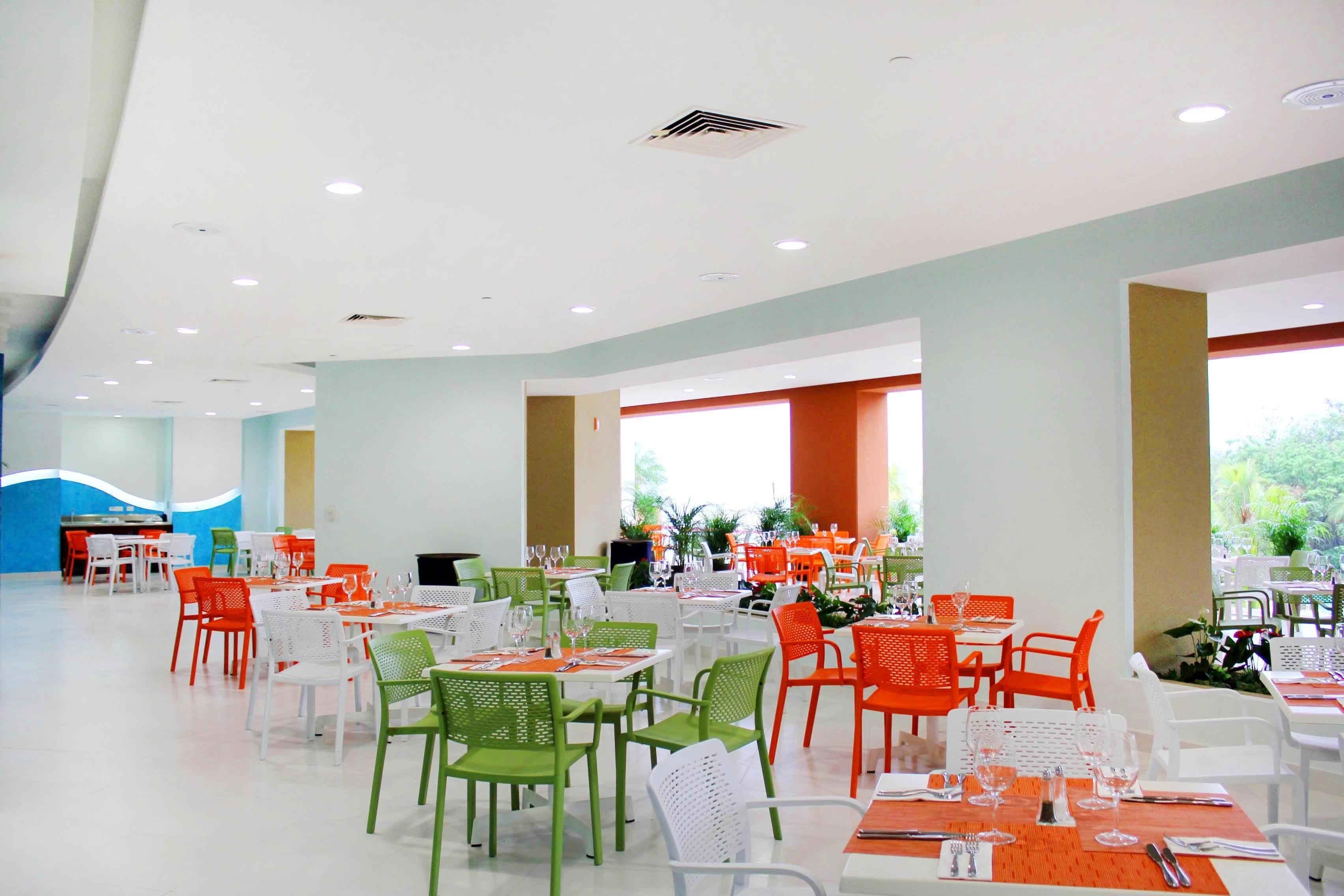 Un amplio comedor interior con varias mesas y sillas, que presenta una combinación de sillas blancas y de colores, y grandes ventanales que permiten la entrada de luz natural.
