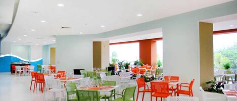 Un amplio comedor interior con varias mesas y sillas, que presenta una combinación de sillas blancas y de colores, y grandes ventanales que permiten la entrada de luz natural.