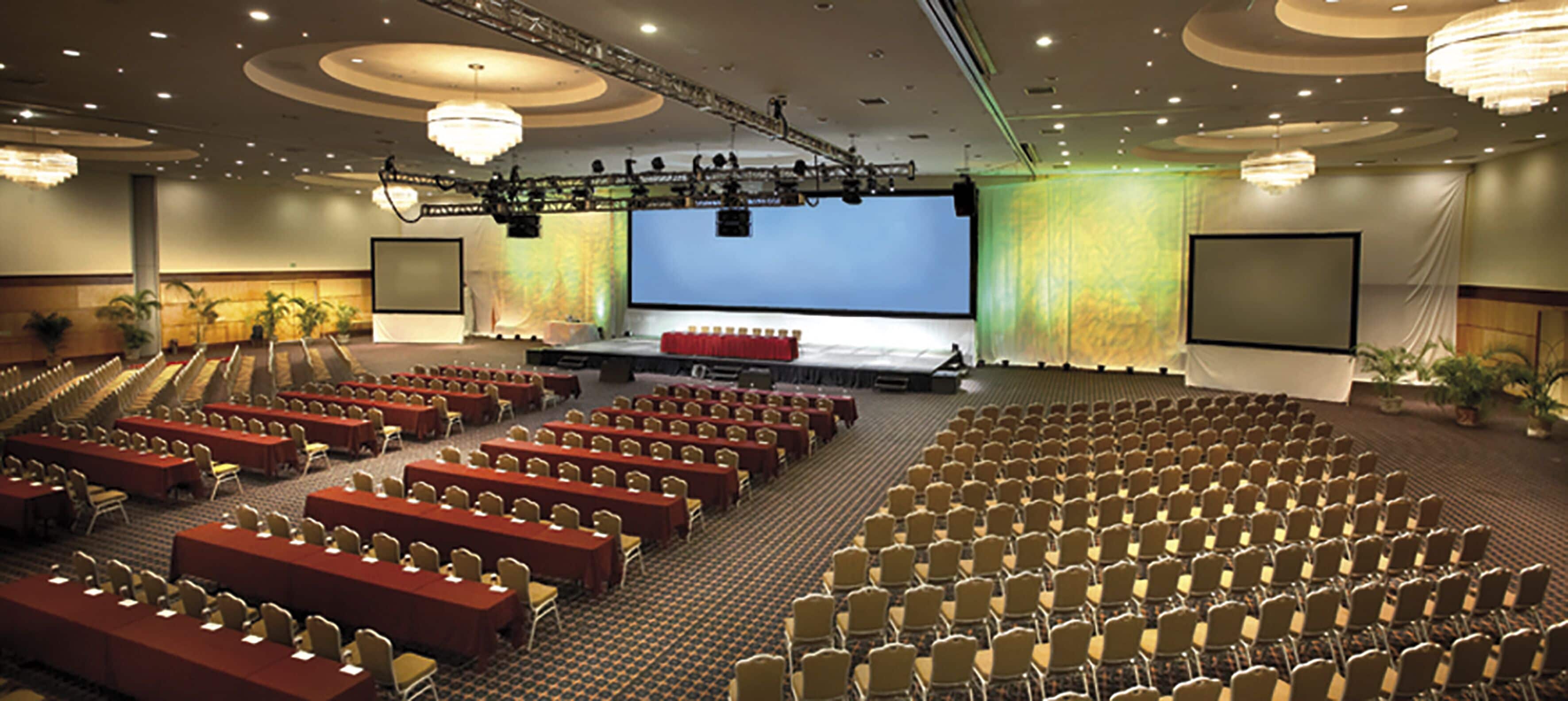 A large conference room with rows of tables and chairs, a stage with a large screen, and chandeliers.