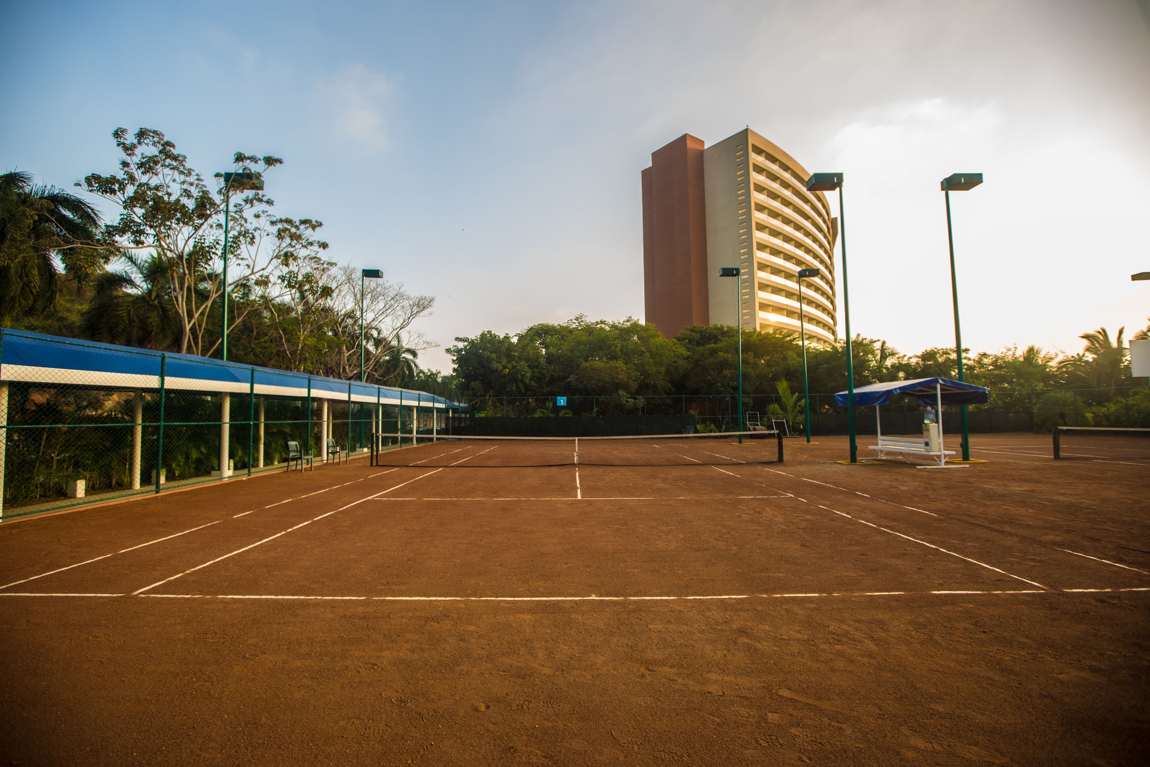 An empty tennis court with a fence, a covered seating area, and a tall building in the background.