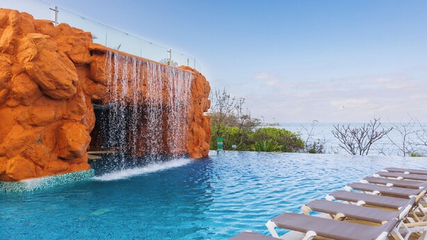 Une piscine avec une cascade artificielle en roche, des chaises longues et une vue sur l’océan.