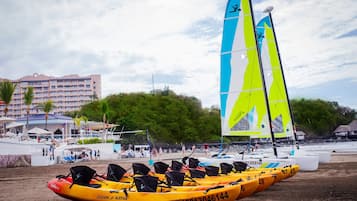 A beach with a row of yellow kayaks lined up, a sailboat with blue and yellow sails, and a hotel building in the background.