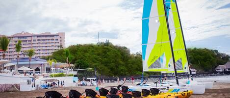 A beach with a row of yellow kayaks lined up, a sailboat with blue and yellow sails, and a hotel building in the background.