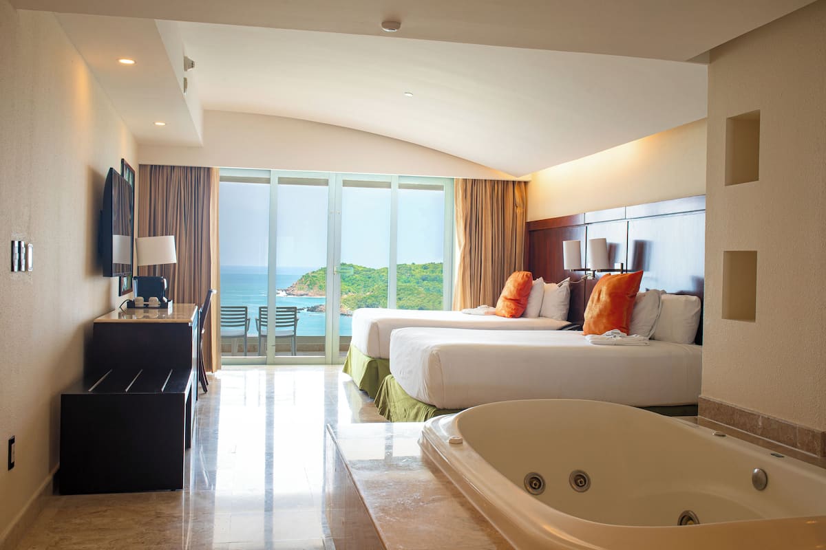 Habitación de hotel con dos camas, una bañera grande, un escritorio con lámpara y vista al mar.