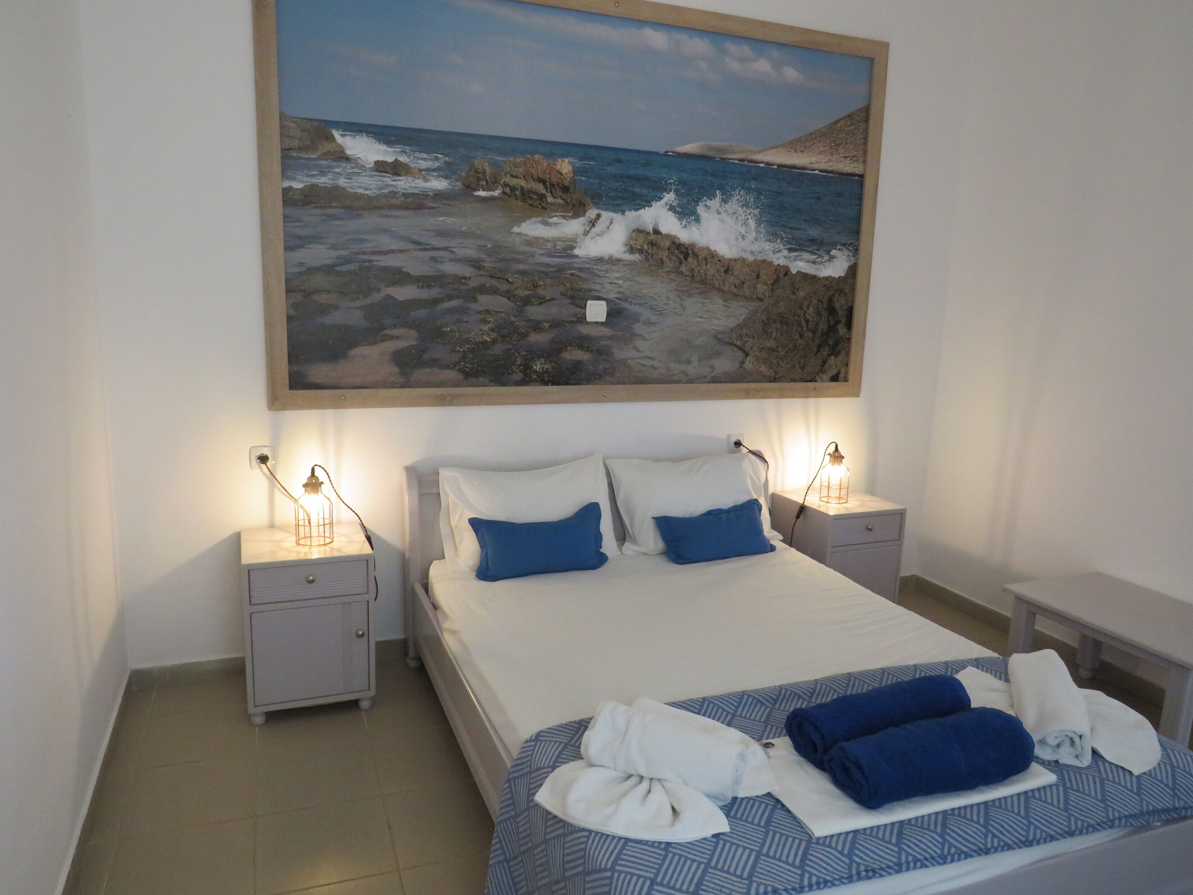 A hotel room with a bed, two bedside tables, a nightstand, a painting of a rocky coastline, and a towel on the bed.