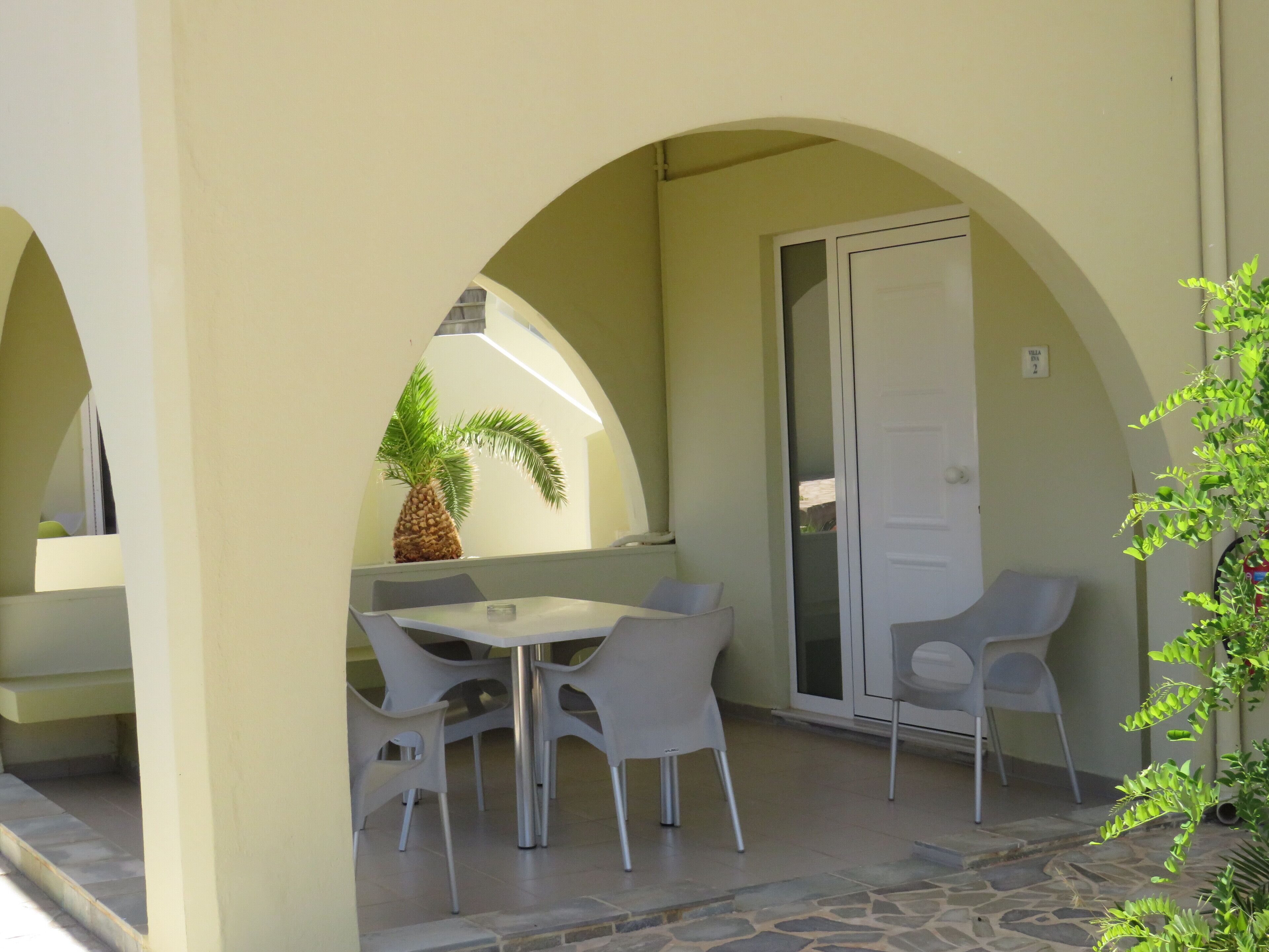 Un espace extérieur abrité, avec une table et des chaises, un palmier et une porte.