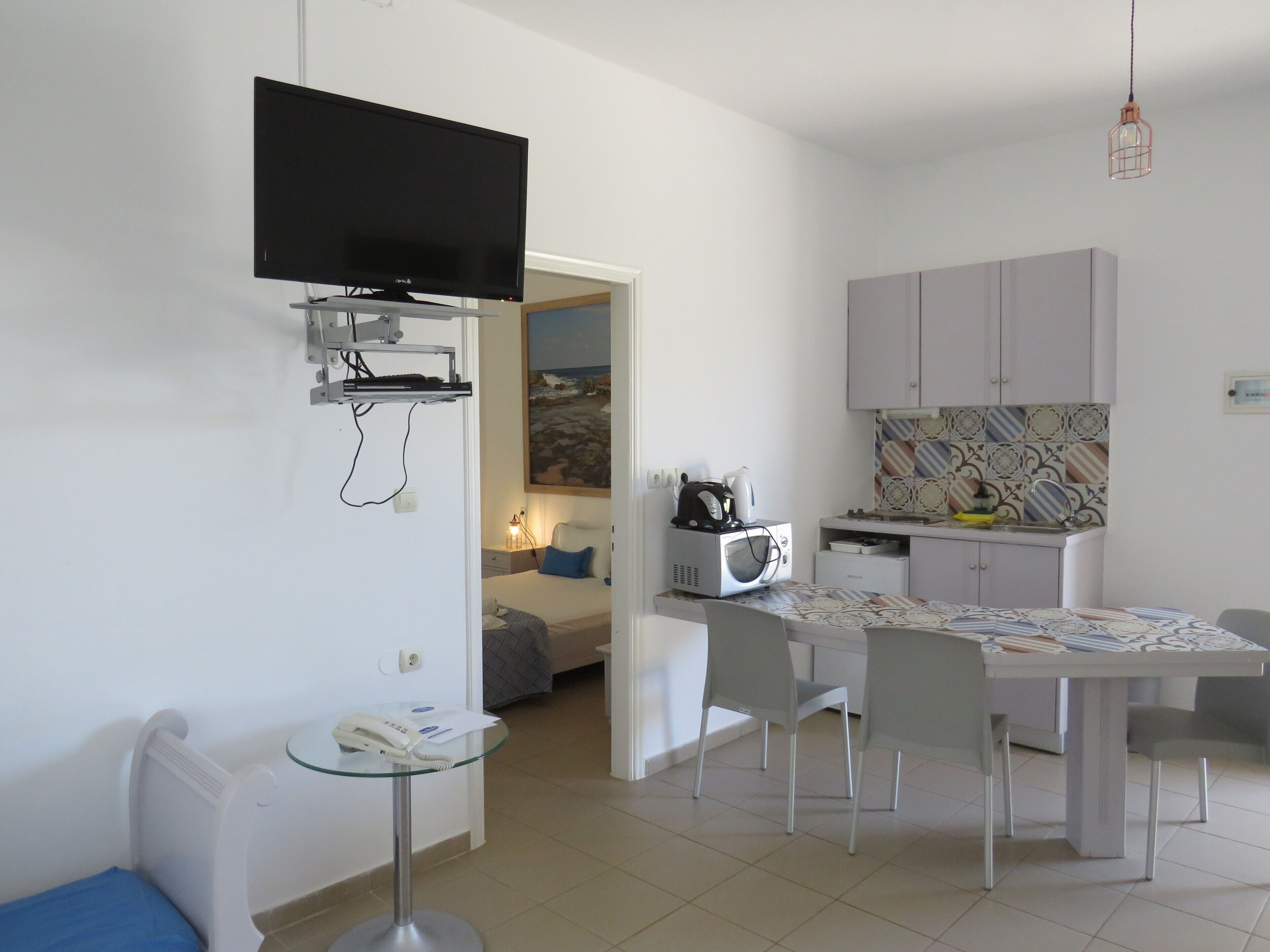 A compact living space with a television mounted on the wall, a small dining table with chairs, a kitchenette with a microwave, and a bed with a view of the outdoors.