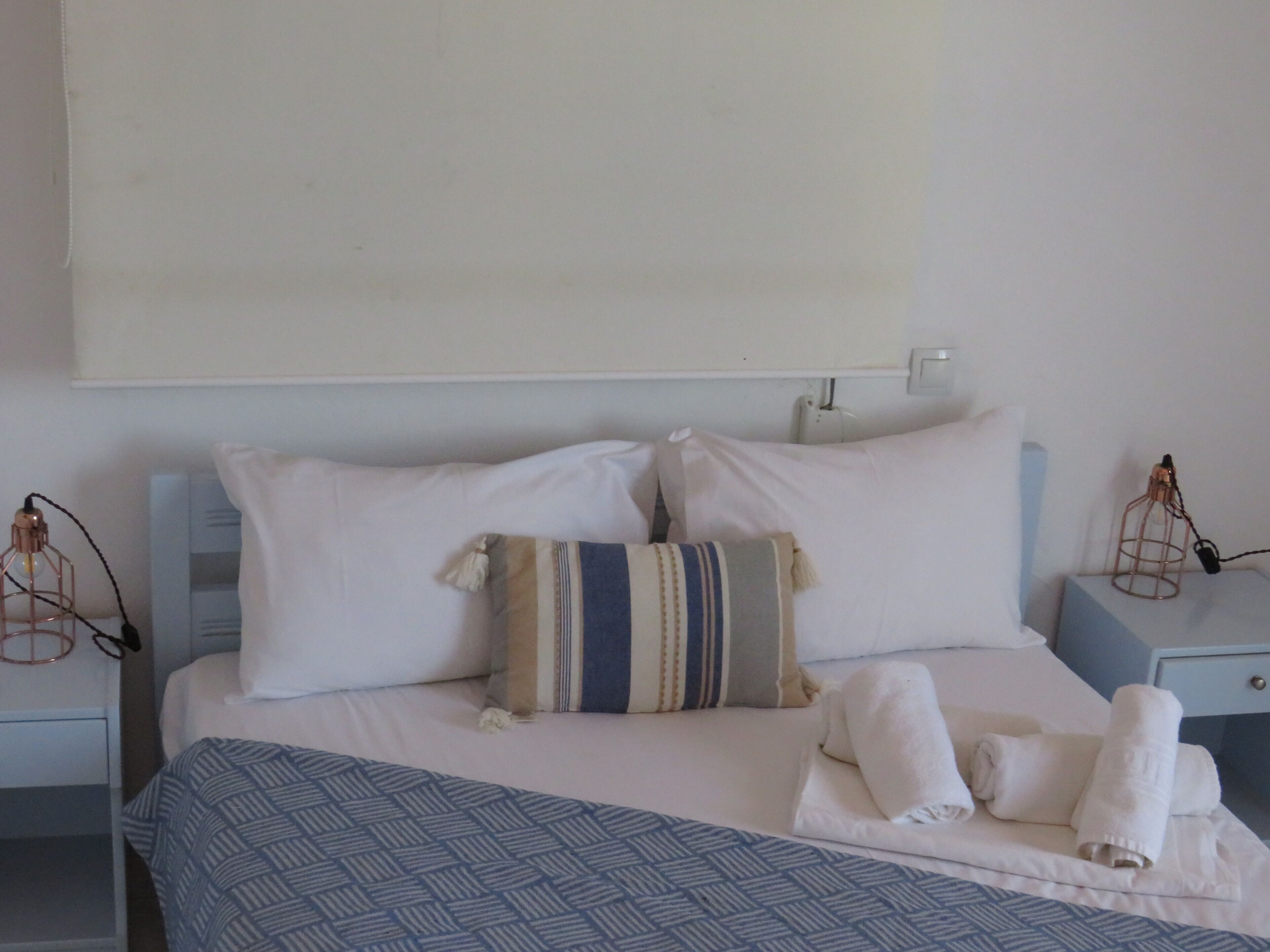 A neatly made bed with white and patterned pillows, a blue patterned bedspread, and rolled towels on the side.