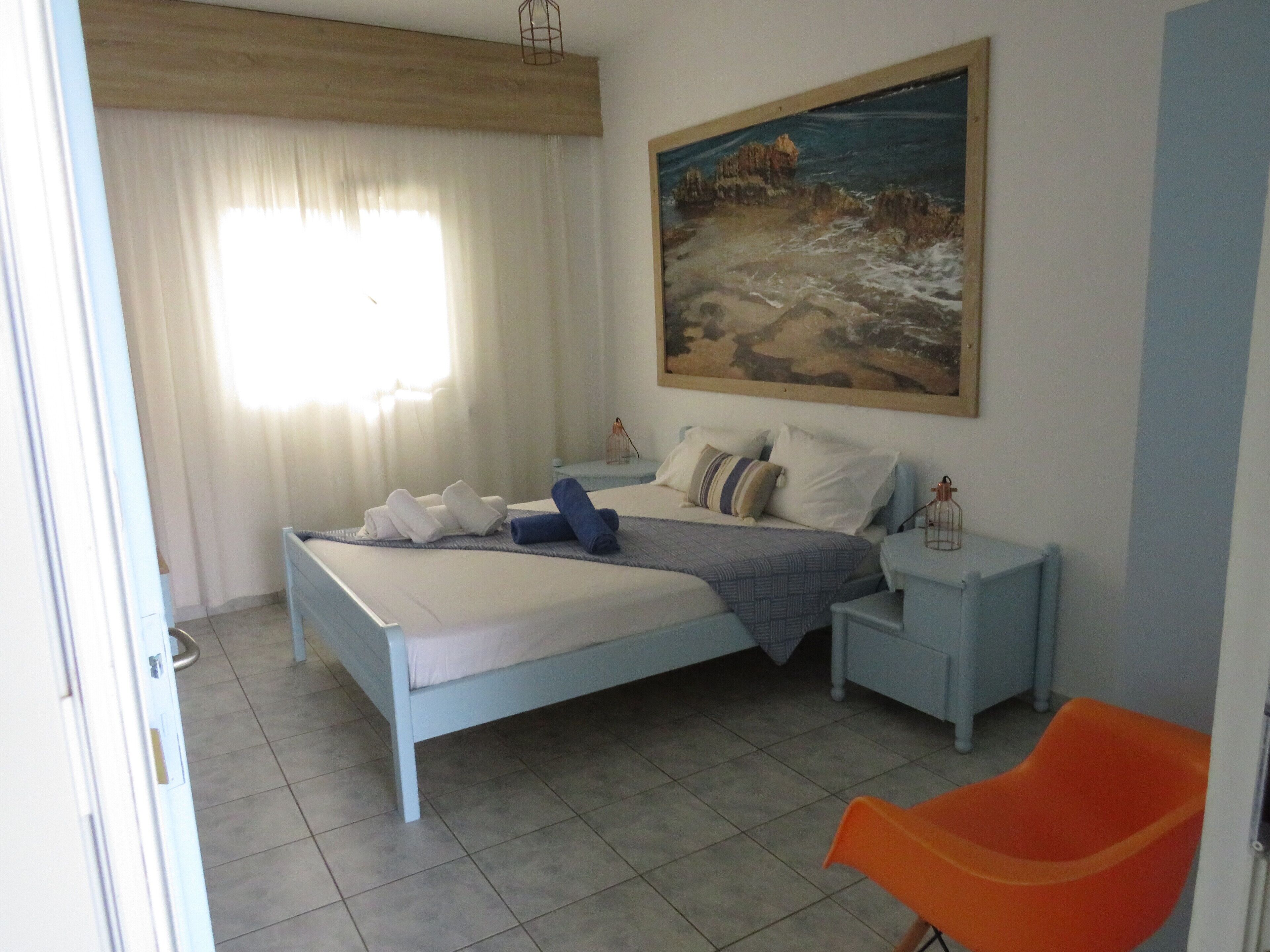 Ein Schlafzimmer mit Bett, Nachttischen, einem Gemälde, einem Sessel und einem Fenster mit Vorhängen.