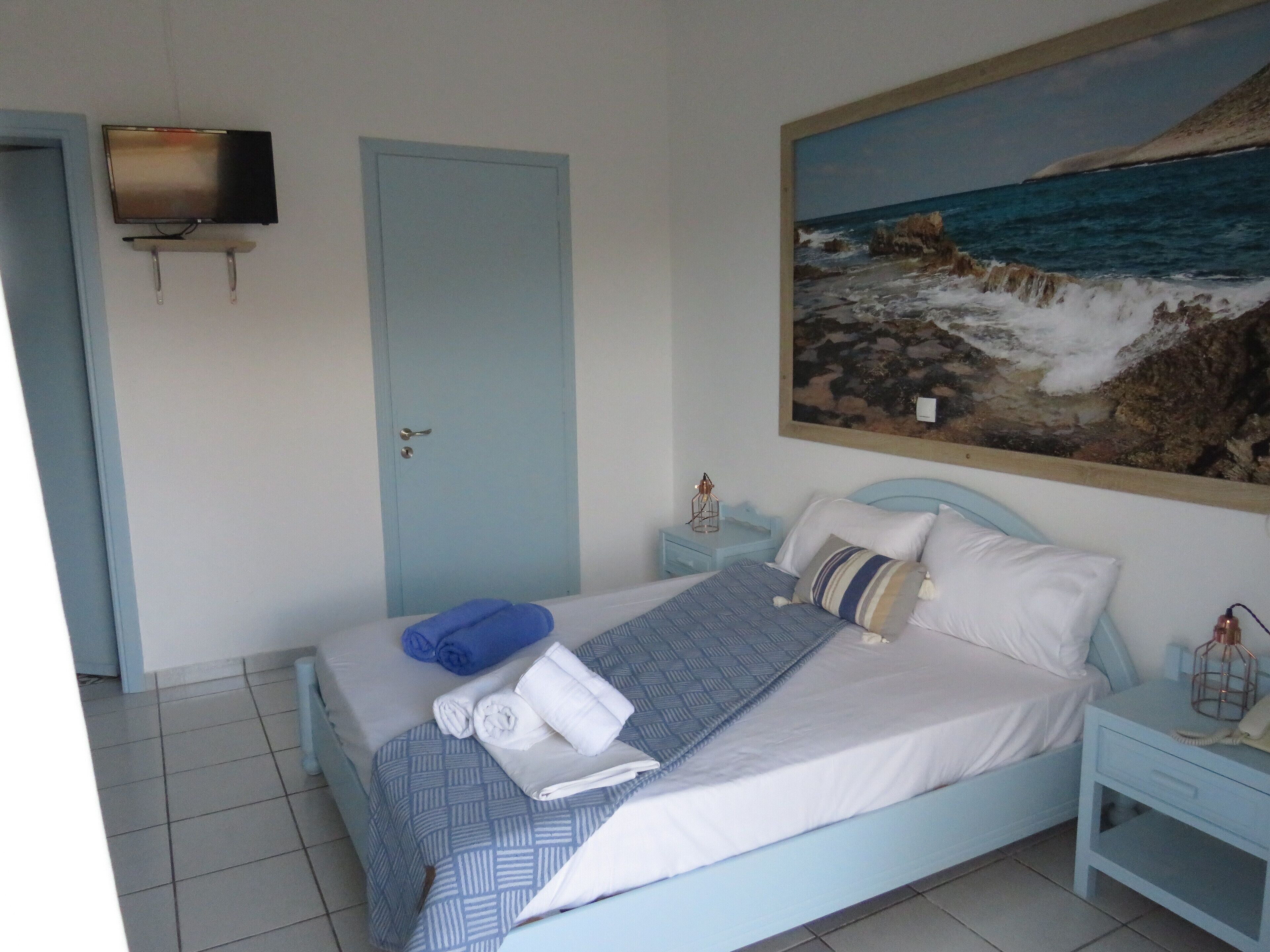 A bedroom with a bed, a nightstand, a TV mounted on the wall, and a painting of a rocky coastline.